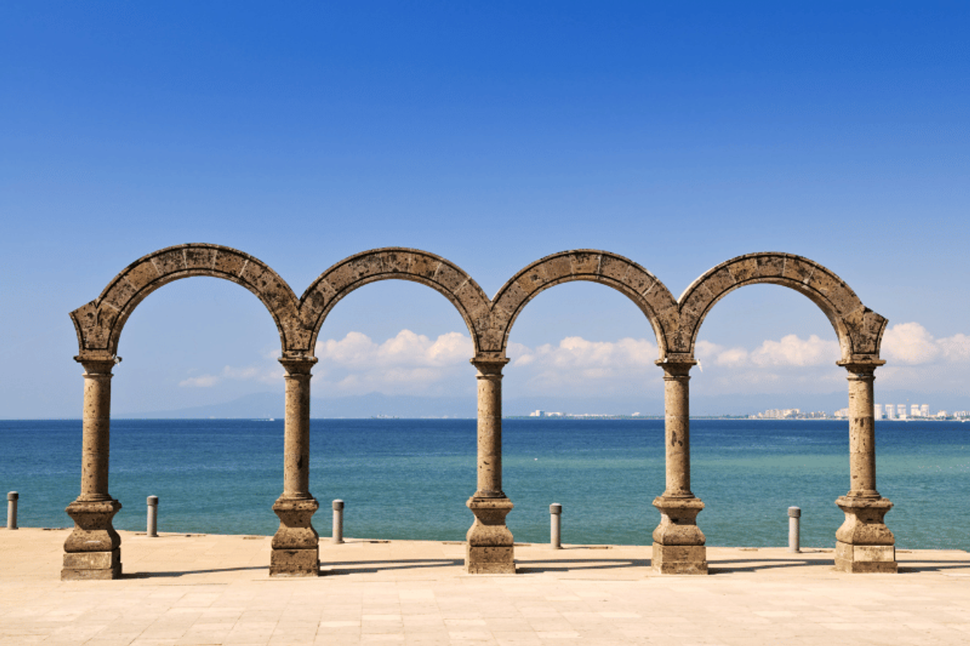 Four stone arches standing in a row along the Malecón in Puerto Vallarta, with the ocean and distant cityscape visible through the arches. The sky is clear and blue, adding to the scenic view.