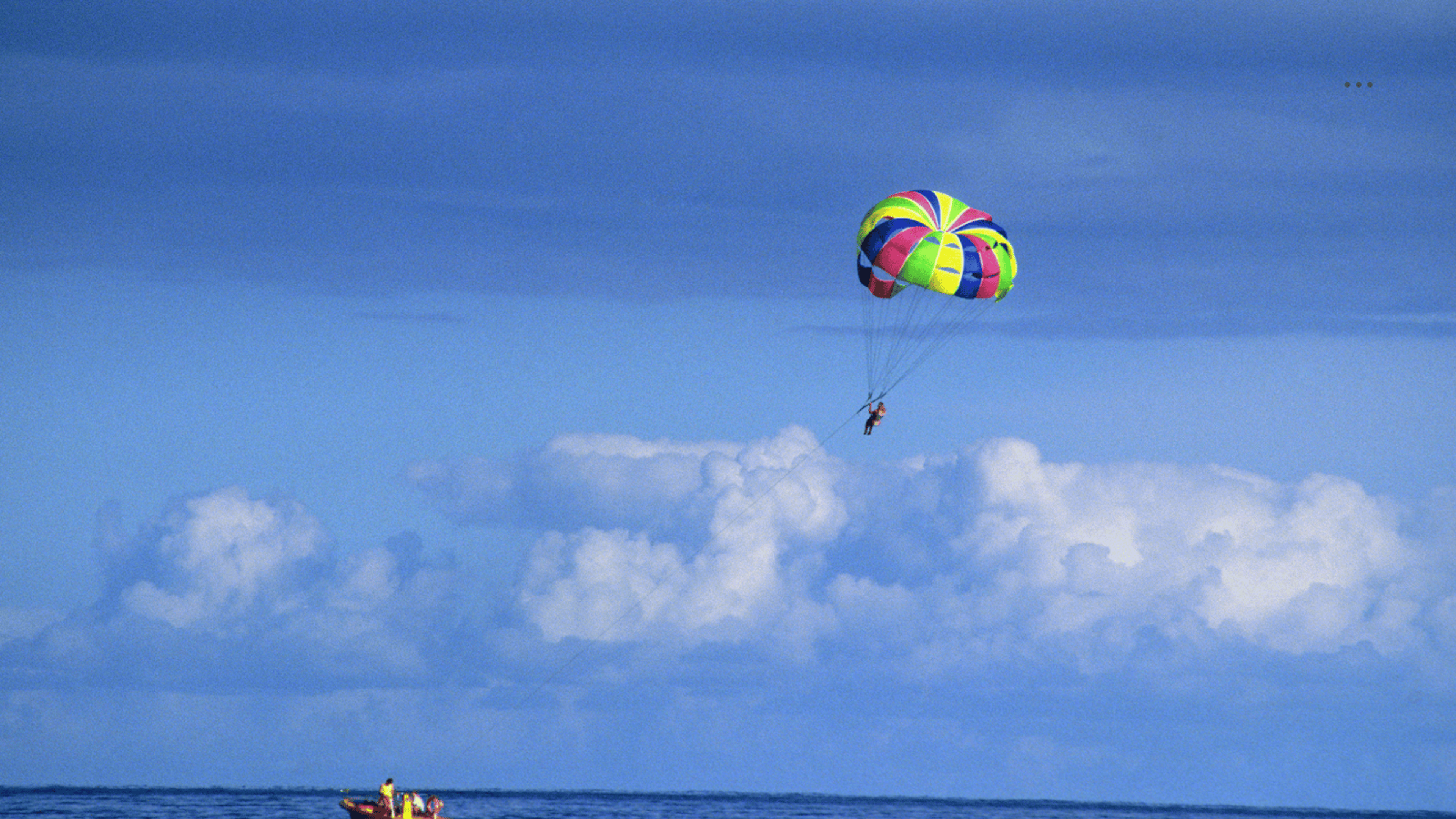 Una persona haciendo parasailing sobre el océano, atada a un paracaídas colorido con un fondo de cielo azul y nubes esponjosas. Un bote es visible abajo, remolcando el parasail.