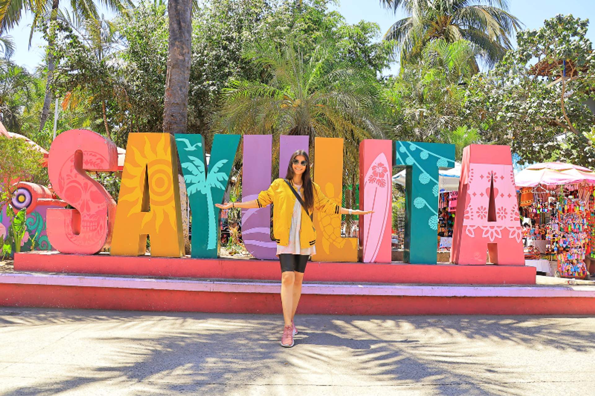 A woman in a yellow jacket poses in front of large, colorful letters spelling "SAYULITA" with palm trees and a market stall in the background.