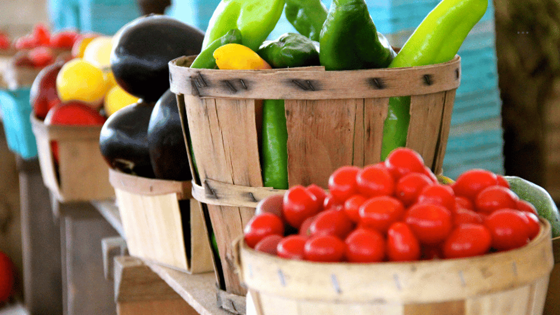 Cestas de madera llenas de verduras frescas en un mercado, incluyendo tomates cherry, pimientos verdes, calabacines amarillos y berenjenas. Los productos son vibrantes y coloridos, exhibidos en un mostrador de madera.