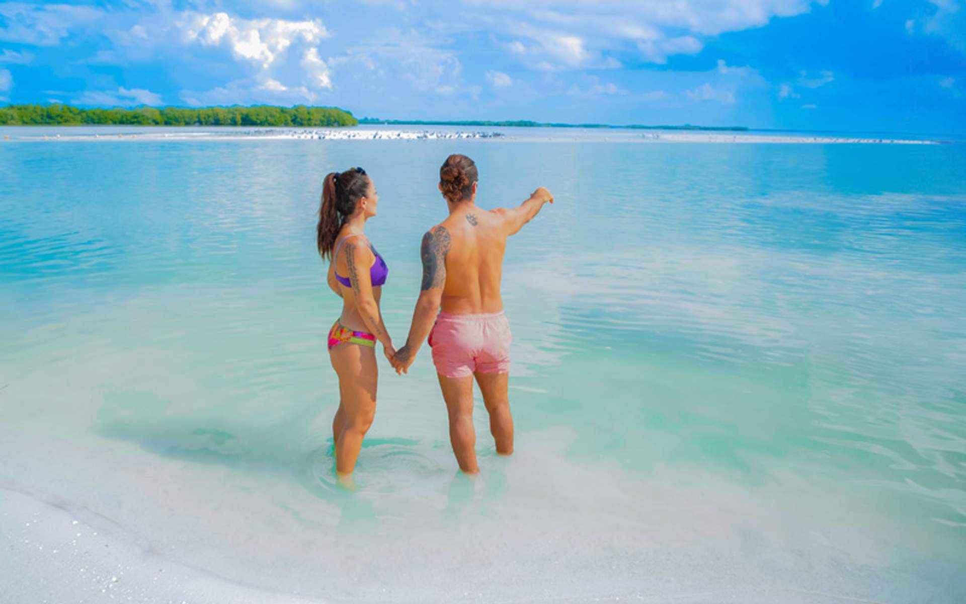 Couple holding hands and walking in shallow beach water, pointing at the horizon.