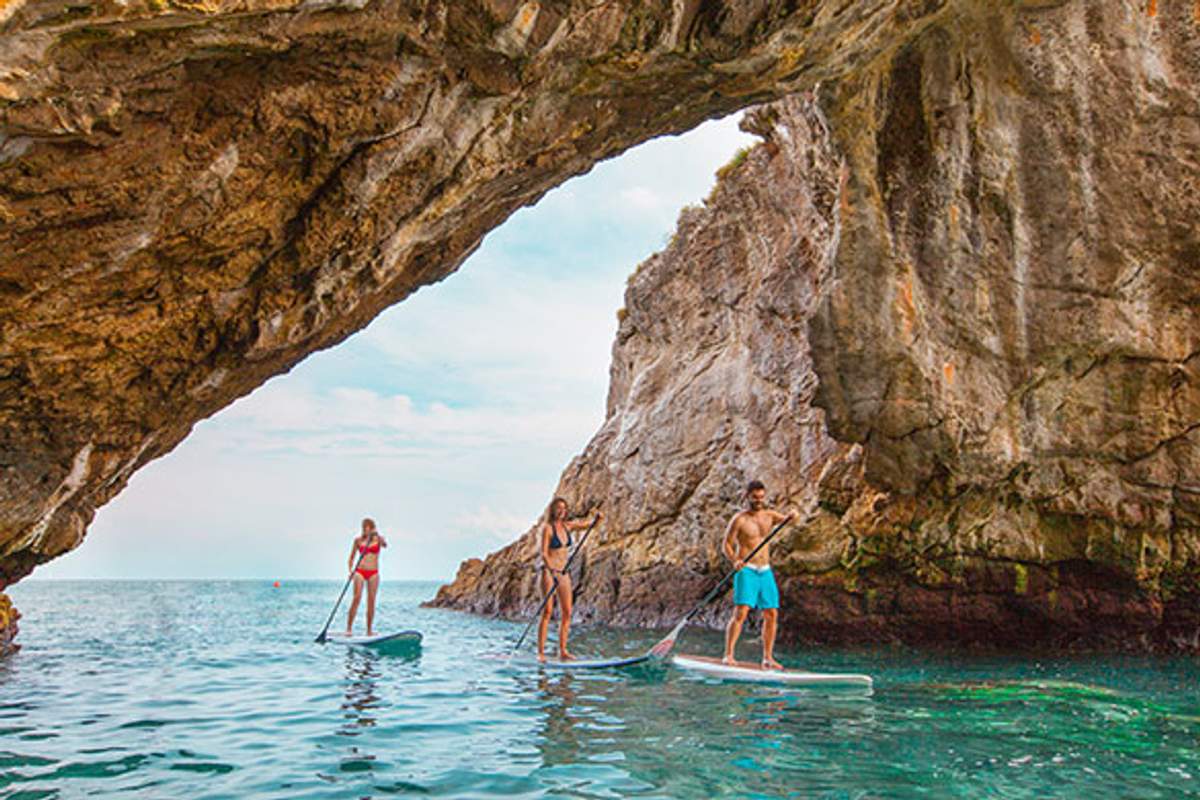 Three people paddleboarding under a large rocky arch in clear blue waters with a view of the open sea.