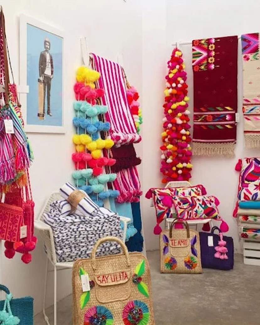 Una exhibición colorida de bolsas, mantas y decoraciones con pompones en una boutique, con textiles vibrantes y una foto enmarcada.