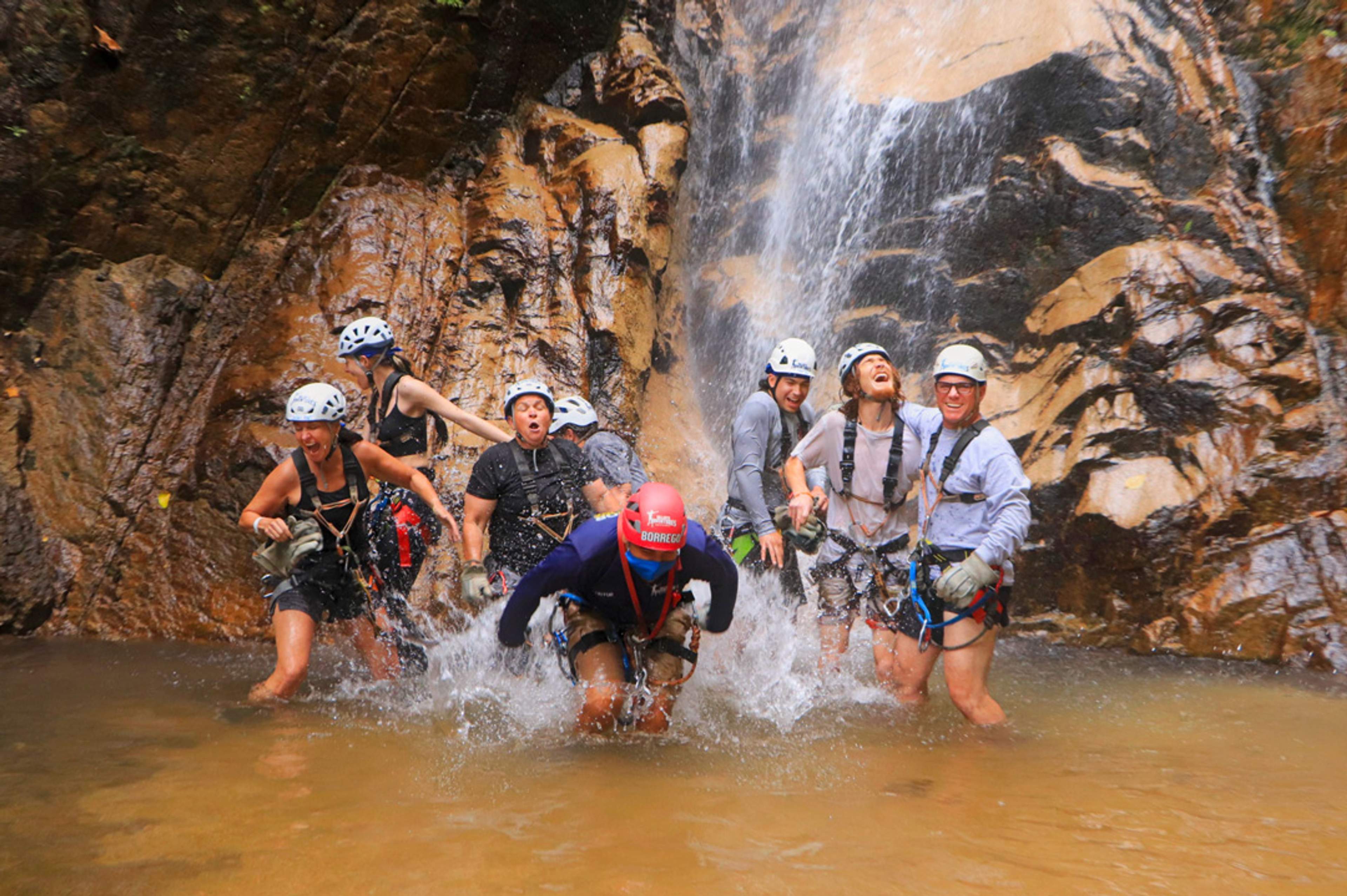 A group of people wearing helmets and harnesses stand in a pool of water at the base of a waterfall, laughing and splashing.