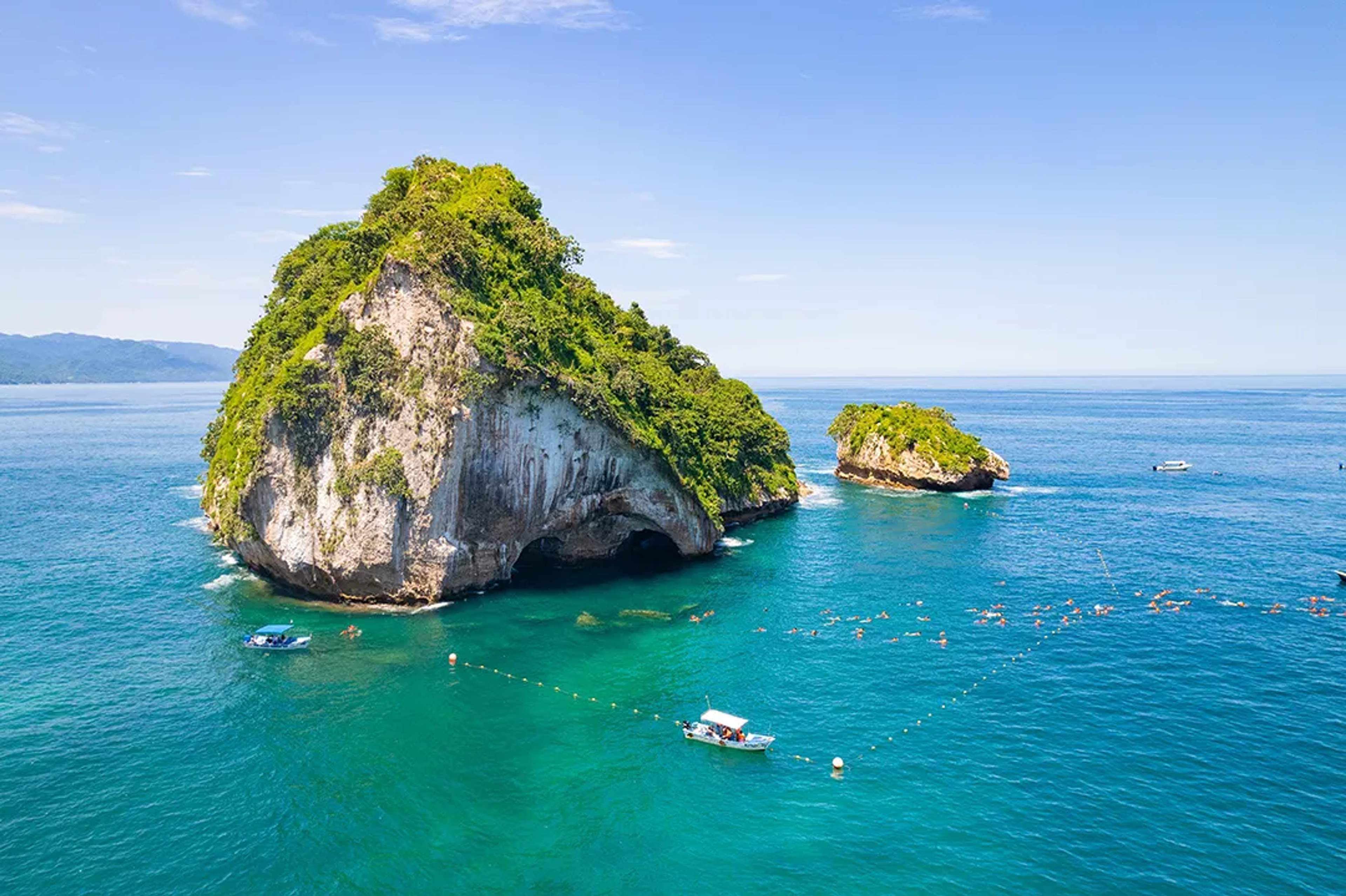 Two lush green rocky islets surrounded by clear blue waters with boats and swimmers at Los Arcos Puerto Vallarta