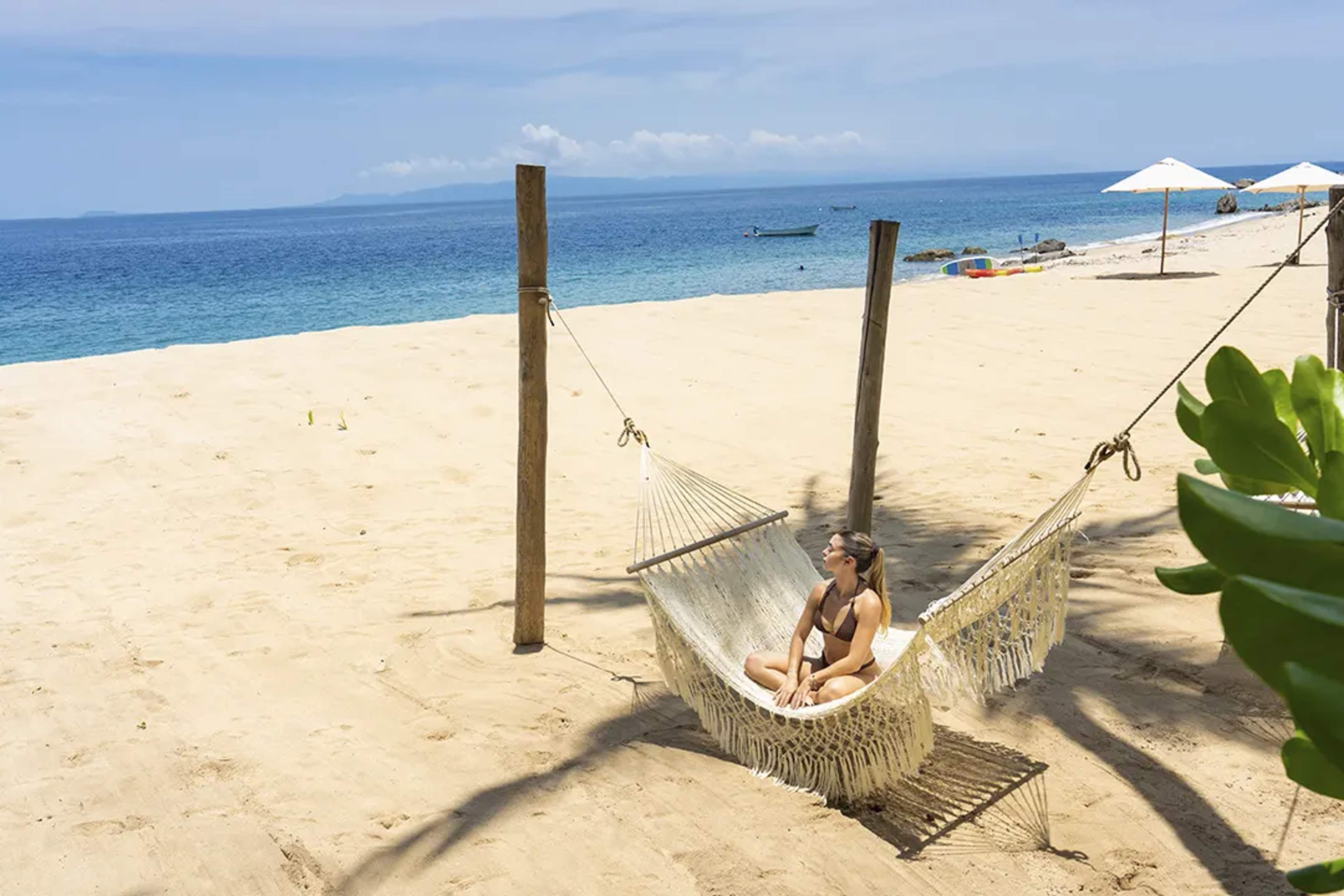 Woman relaxing in a hammock on a sandy beach with a view of the ocean and distant mountains under a clear sky.