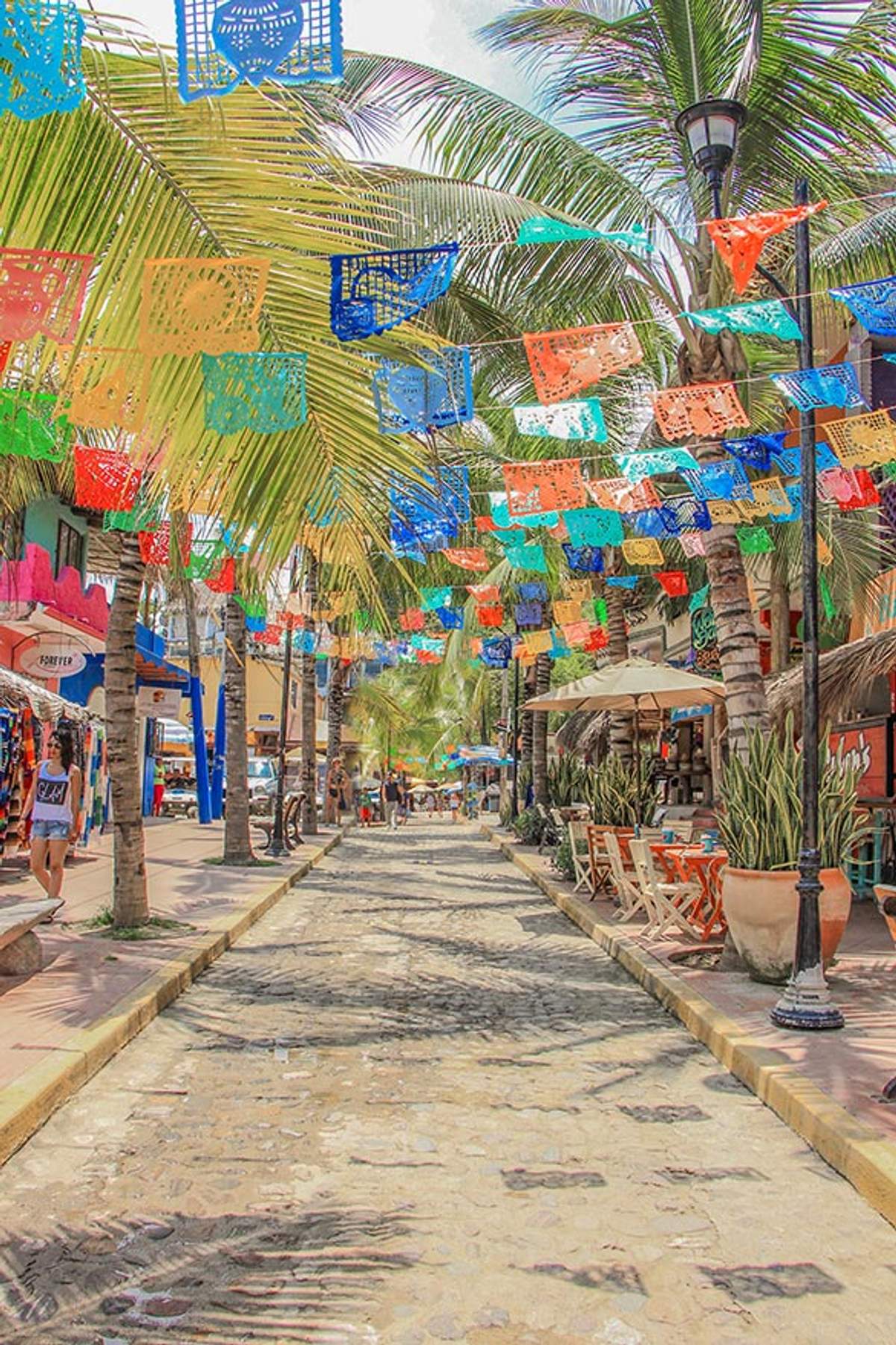 Una calle colorida en Sayulita decorada con vibrantes banderas de papel picado colgando arriba, bordeada de palmeras, tiendas y asientos al aire libre.