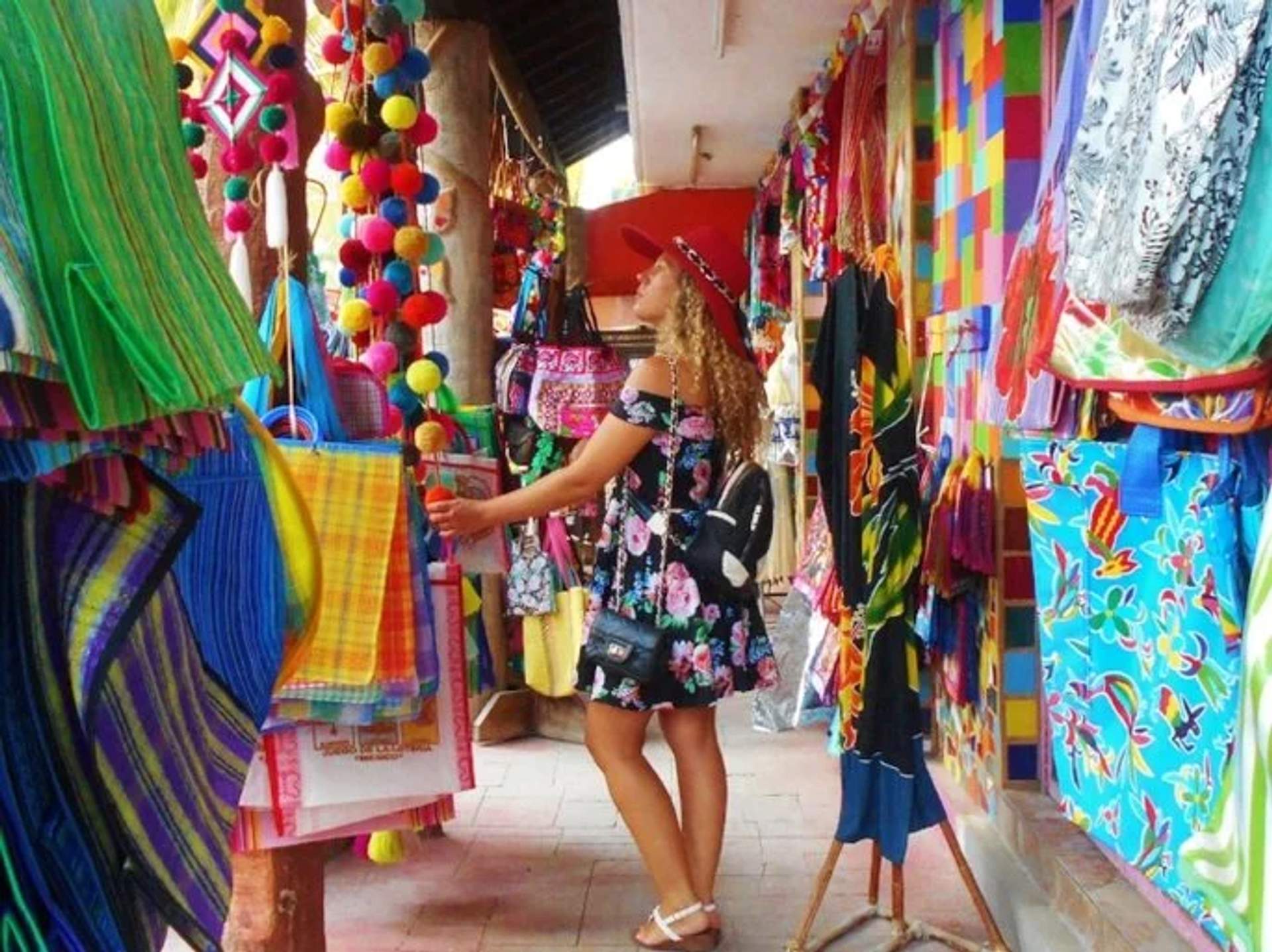 A woman in a floral dress and hat shopping in a colorful Mexican market filled with vibrant textiles and decorations.