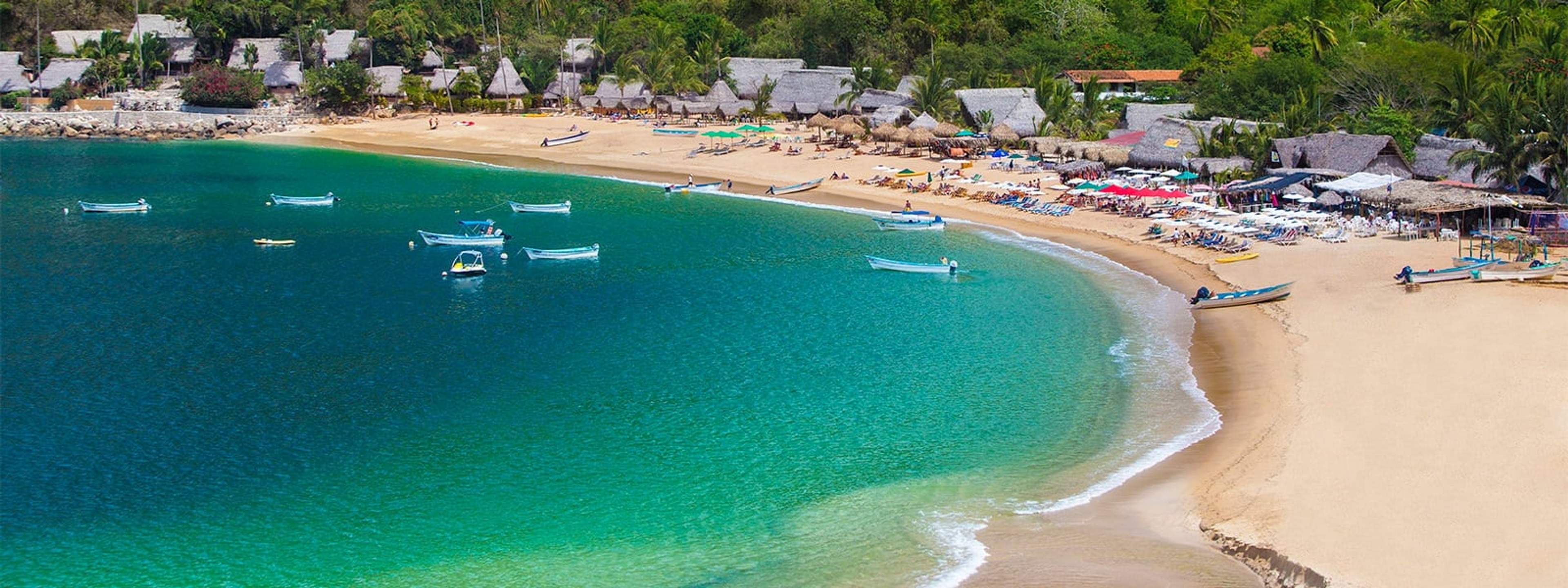 A beautiful beach with golden sand and turquoise water, lined with small boats and thatched-roof huts in the background, along with beachgoers lounging under umbrellas.