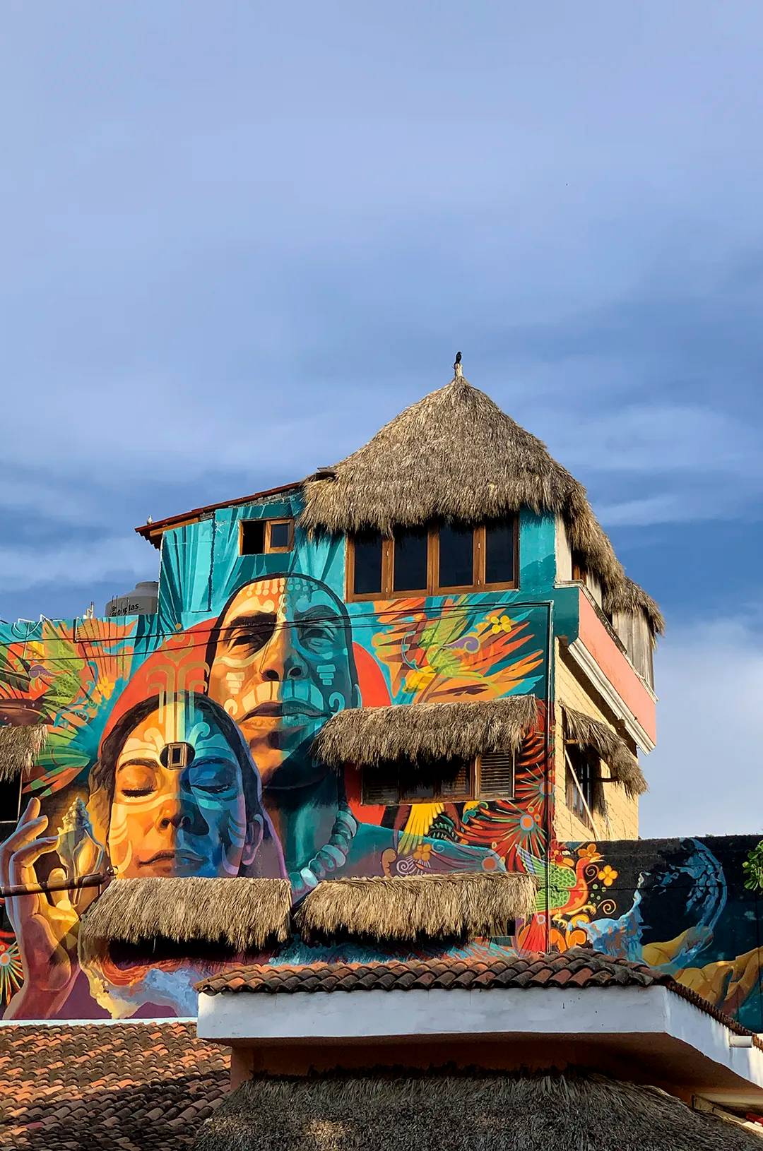 Mural colorido de rostros en un edificio con techos de paja en Sayulita.