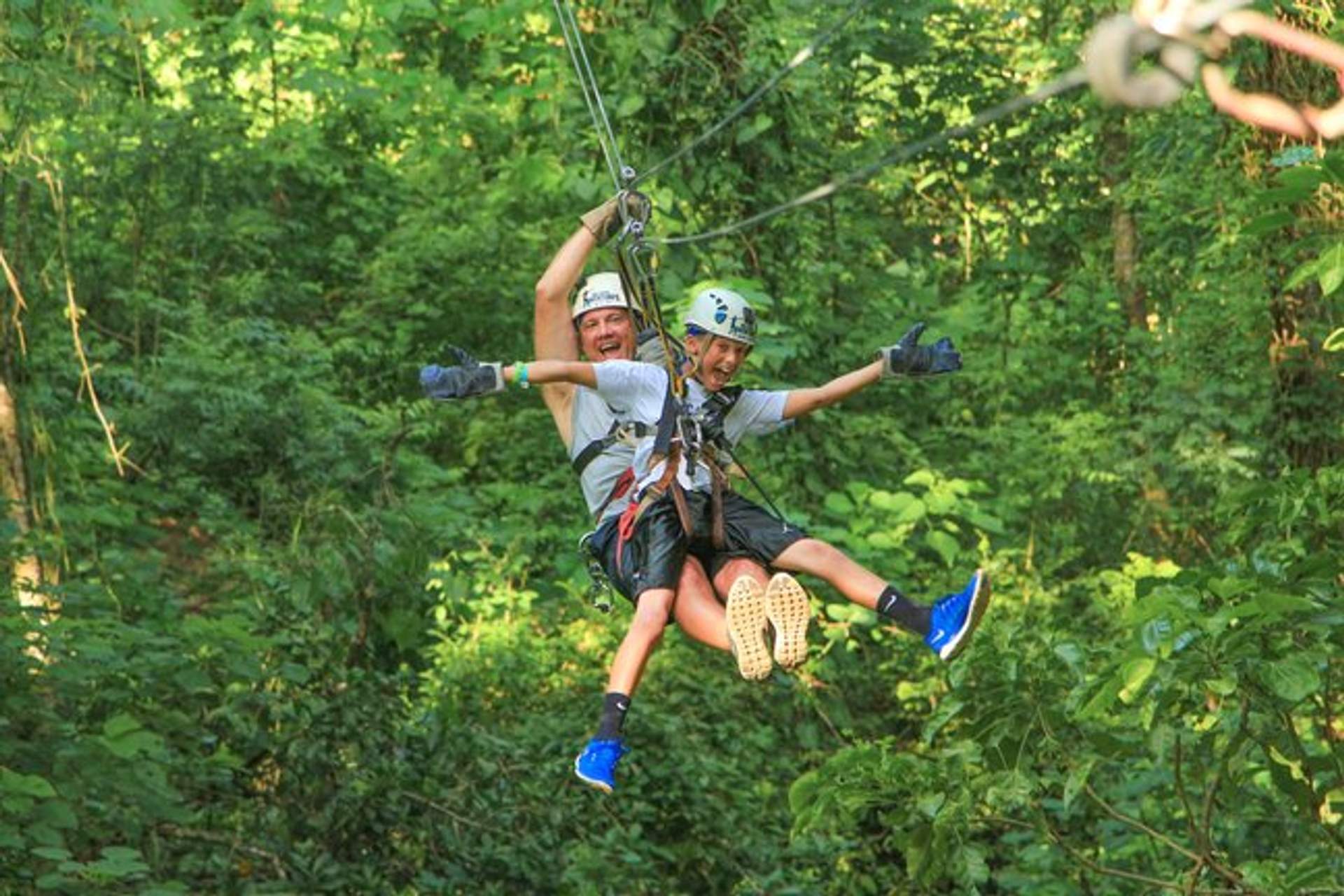 A father and son enjoy a thrilling zipline adventure through a lush forest, both smiling and excited.