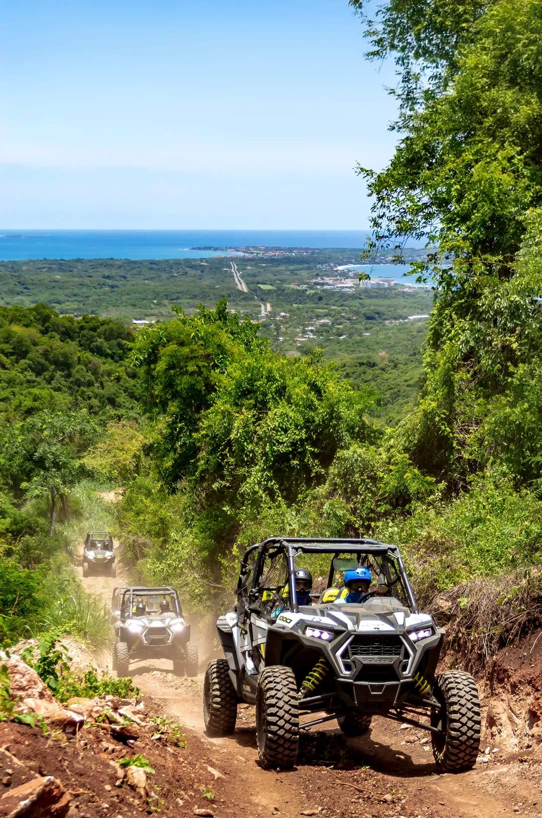 ATVs driving through a lush jungle trail with an ocean view in the background.