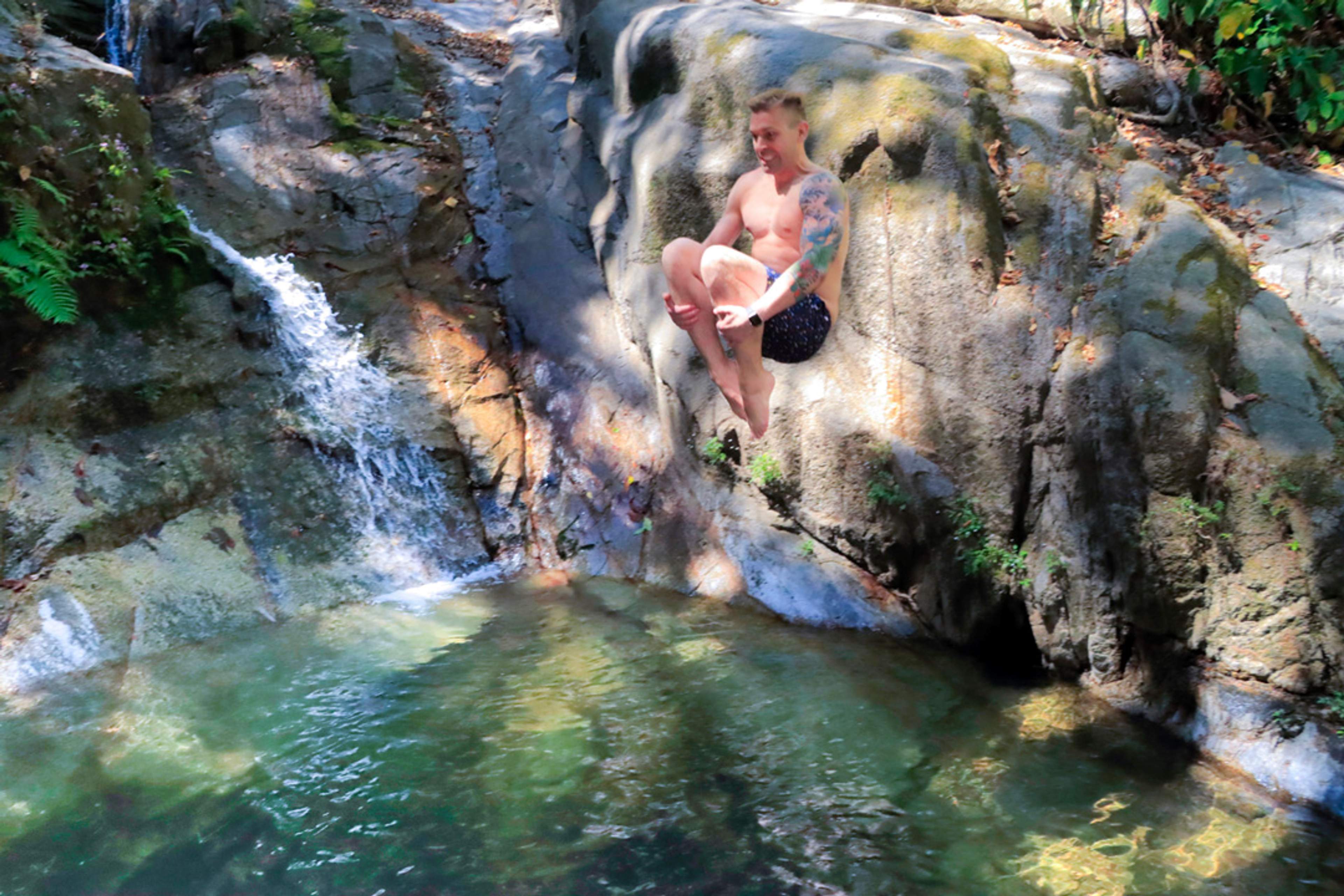 A shirtless man with tattoos jumps into a clear pool of water from a rocky ledge near a small waterfall in a forested area.