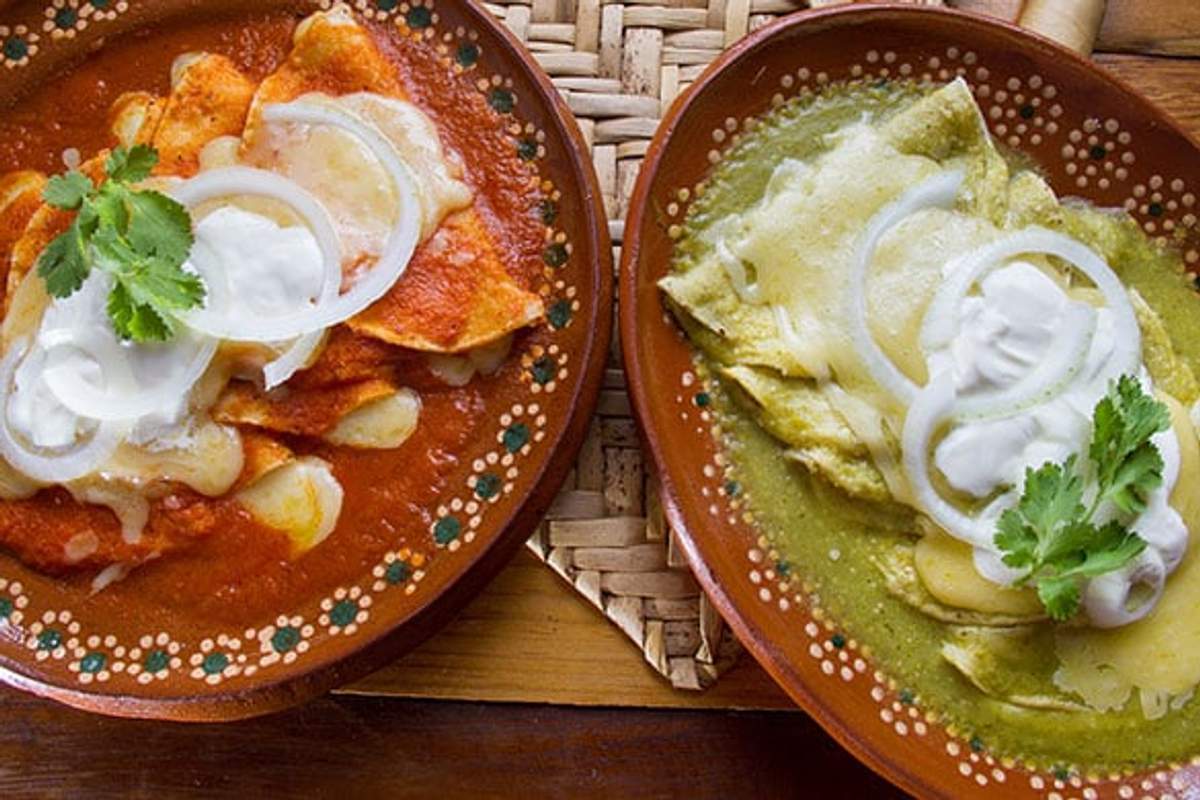 Dos platos de enchiladas, uno con salsa roja y el otro con salsa verde, adornados con cebolla, crema y cilantro.