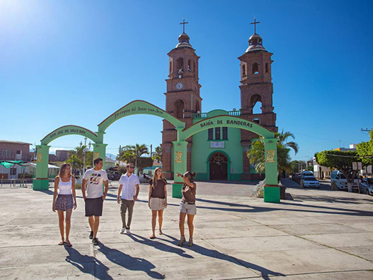 Five people walking in front of a green arch and a church with twin towers under a clear blue sky.