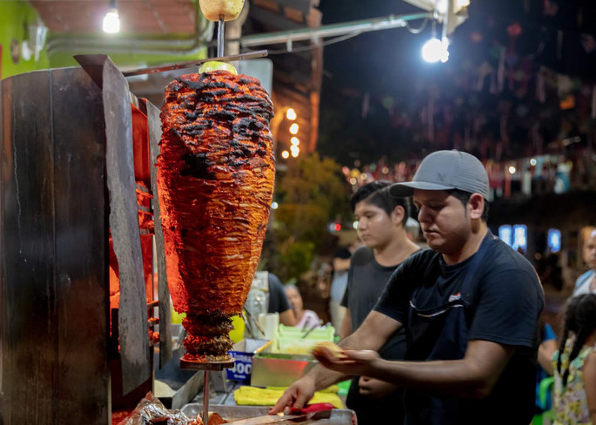 A street vendor prepares tacos al pastor from a large, vertical spit at a bustling night market in Mexico.