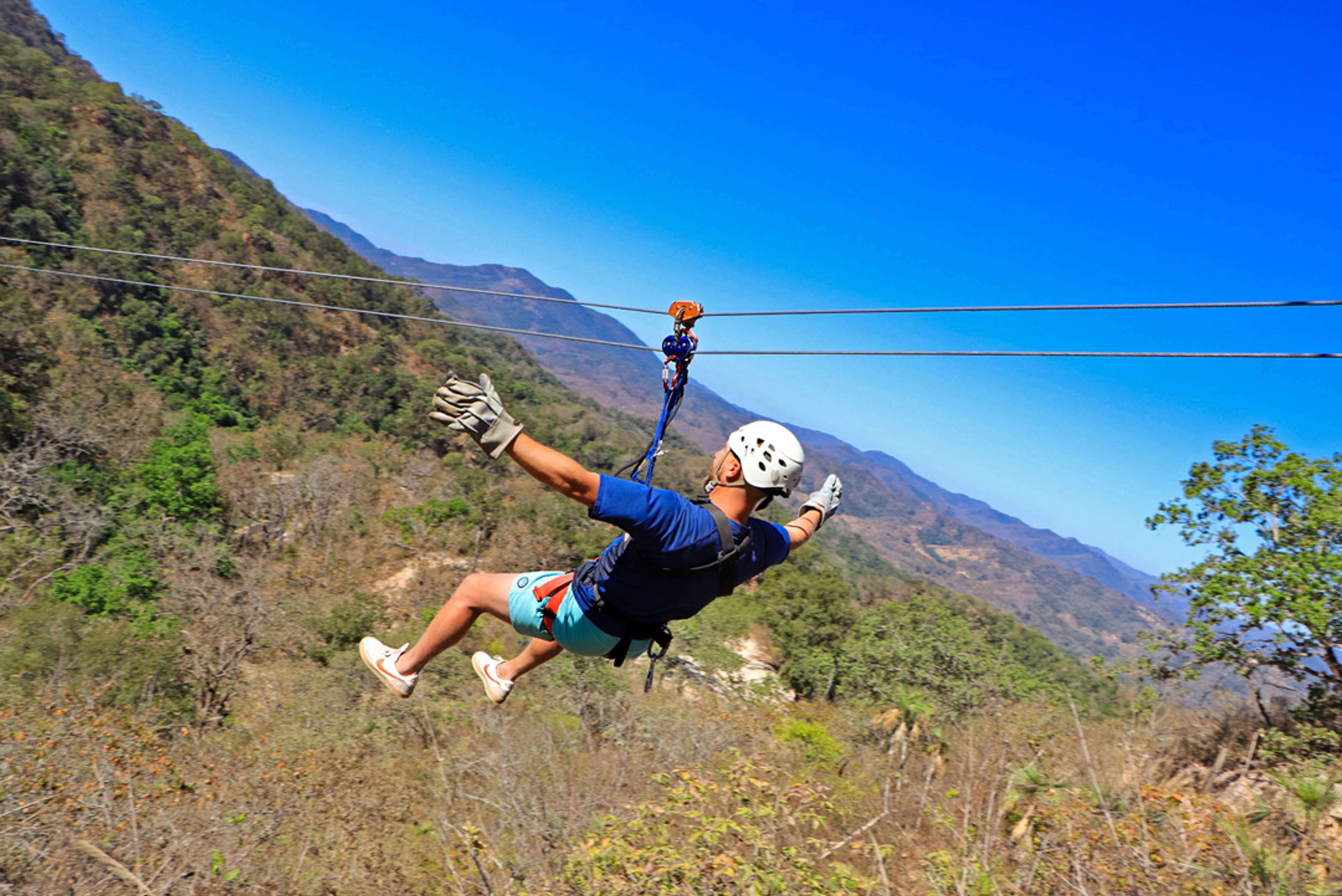 Una persona con casco y guantes monta una tirolesa sobre un valle boscoso con montañas al fondo, extendiendo los brazos a los lados.