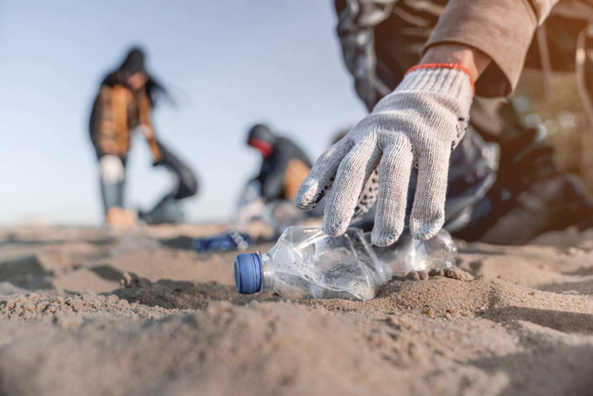 Voluntarios con guantes recogen botellas de plástico y basura de una playa de arena, participando en una jornada de limpieza.