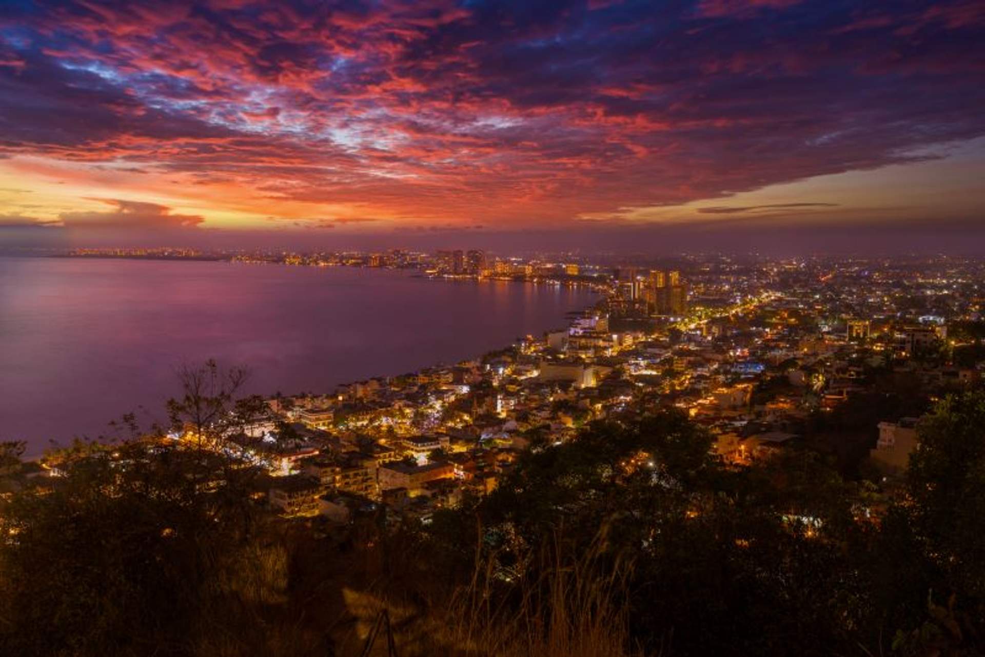 A vibrant cityscape of Puerto Vallarta, Mexico, at sunset with a colorful sky filled with purple, pink, and orange hues. The city's lights are beginning to illuminate as night falls, reflecting on the tranquil waters of Banderas Bay.