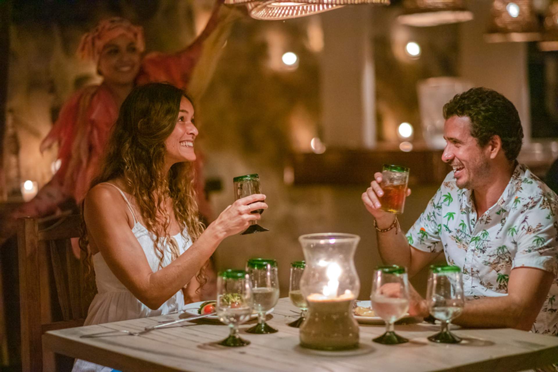 A couple enjoys a candlelit dinner, toasting with drinks, while a performer in the background adds to the festive atmosphere.
