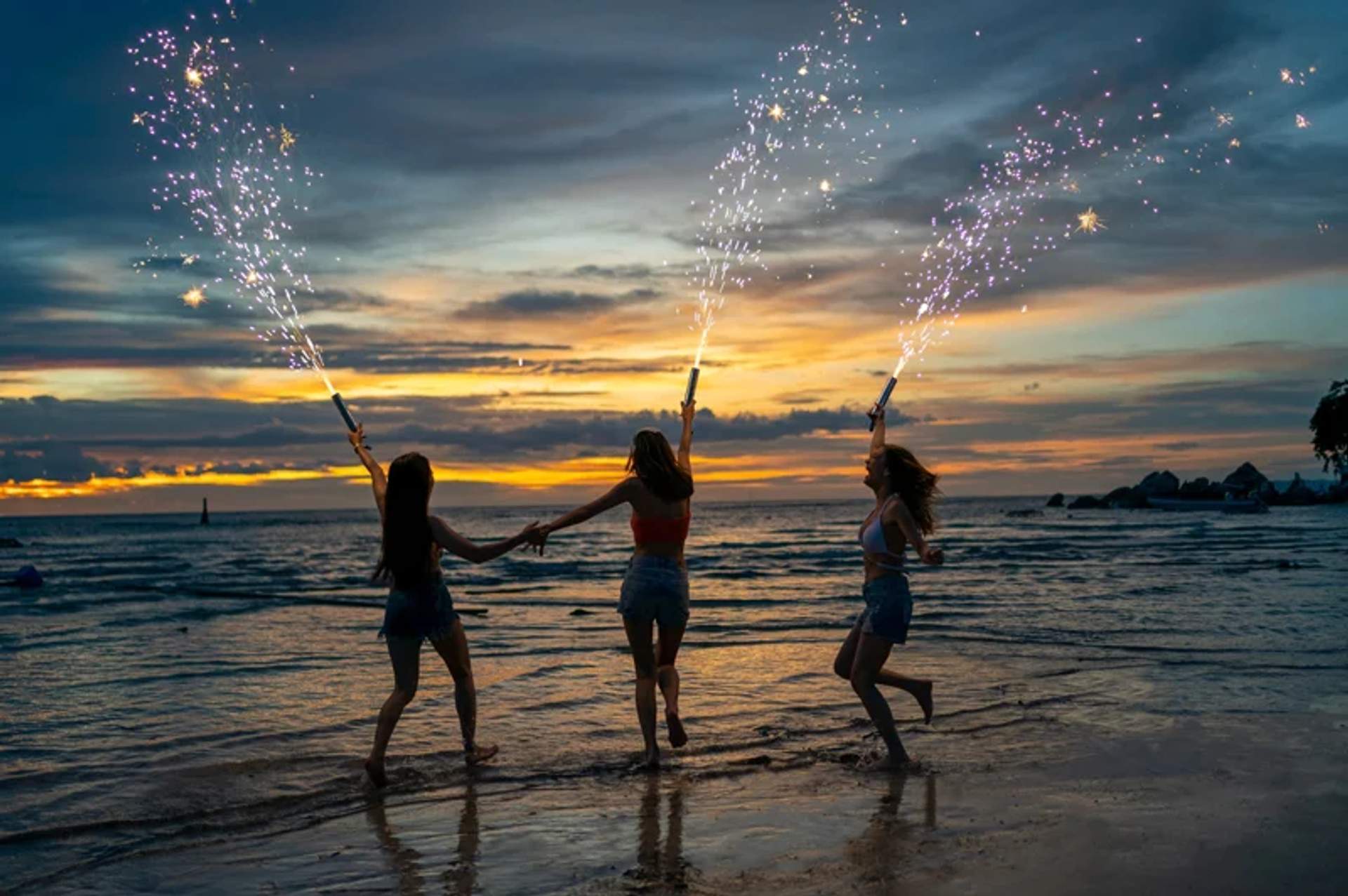Tres mujeres sosteniendo bengalas y bailando en la playa al atardecer. El cielo está lleno de colores vibrantes, y los reflejos en el agua añaden a la atmósfera festiva y alegre.