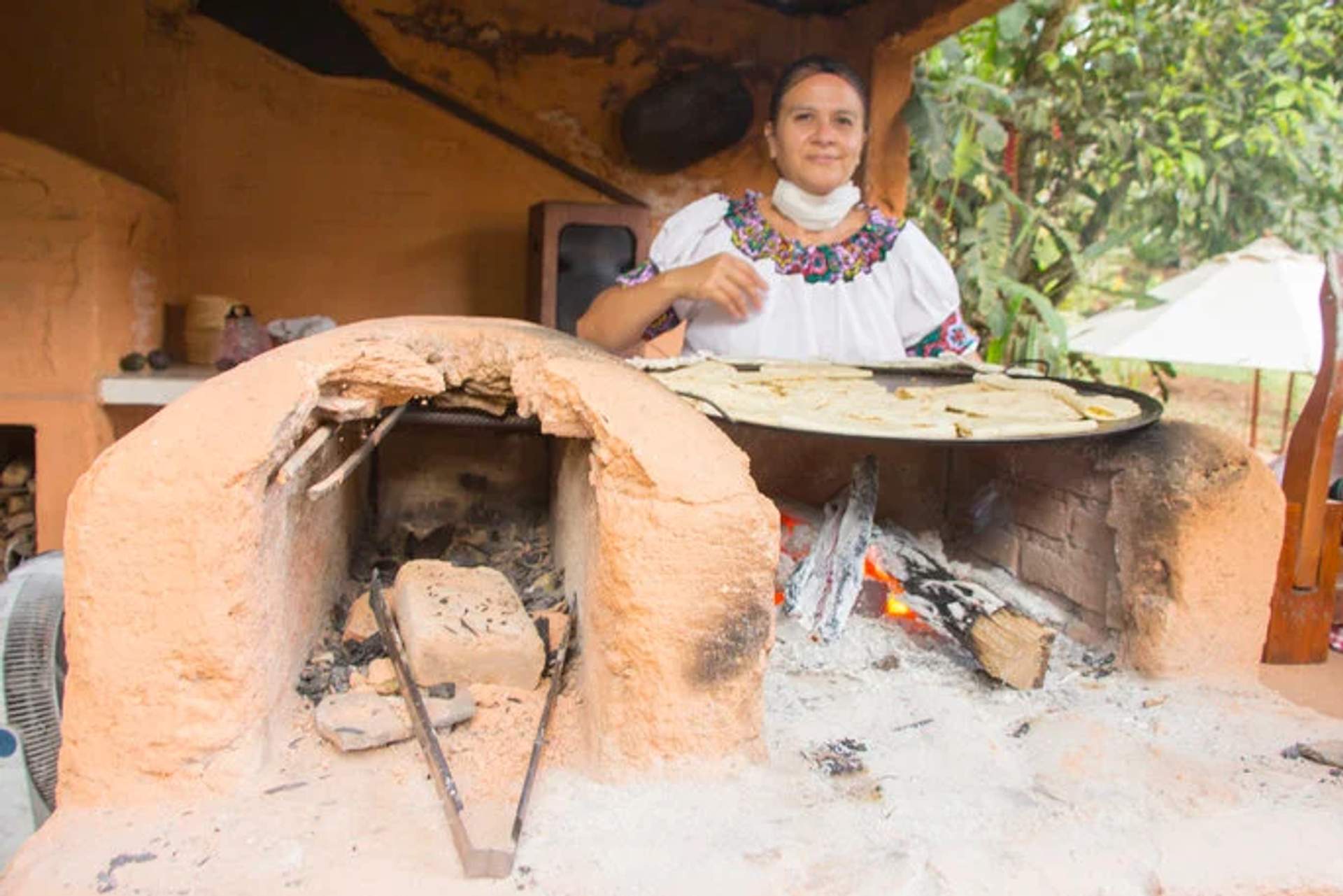 Una mujer con atuendo tradicional cocina tortillas en un rústico horno de leña, mostrando la cocina tradicional mexicana.