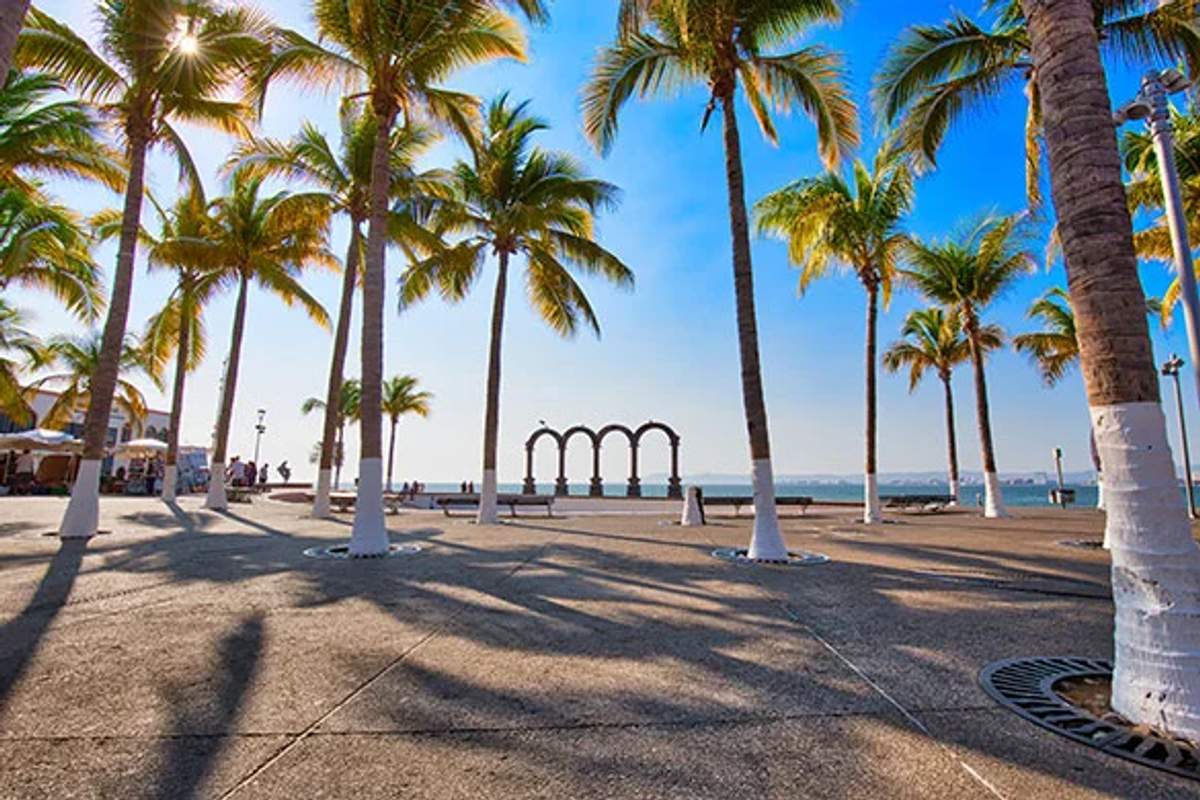Palmeras bordean el paseo que conduce a los icónicos arcos del Malecón de Puerto Vallarta, con el océano de fondo.