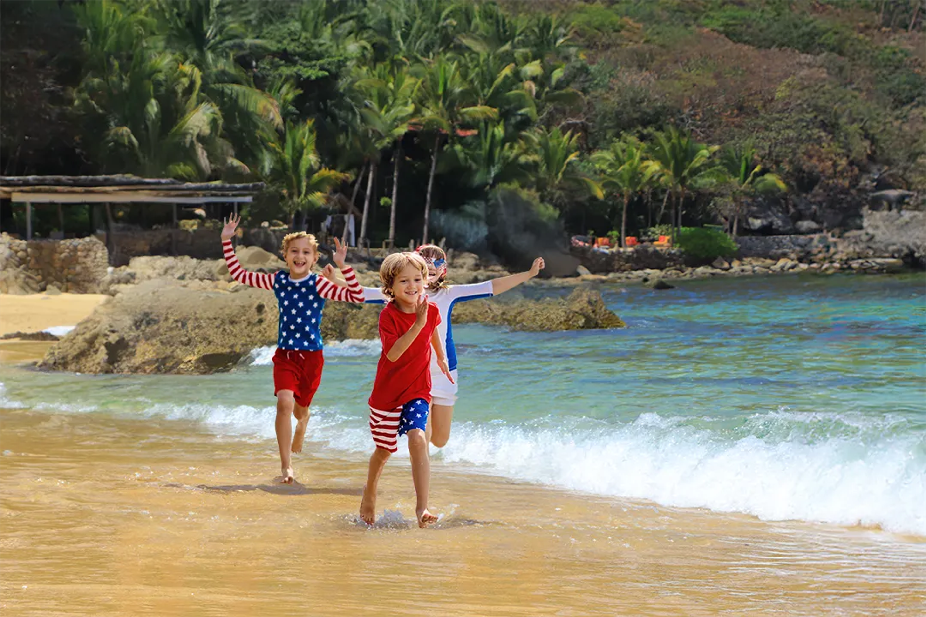 Three children wearing patriotic-themed clothing run along the beach at Las Caletas, Puerto Vallarta, Mexico.