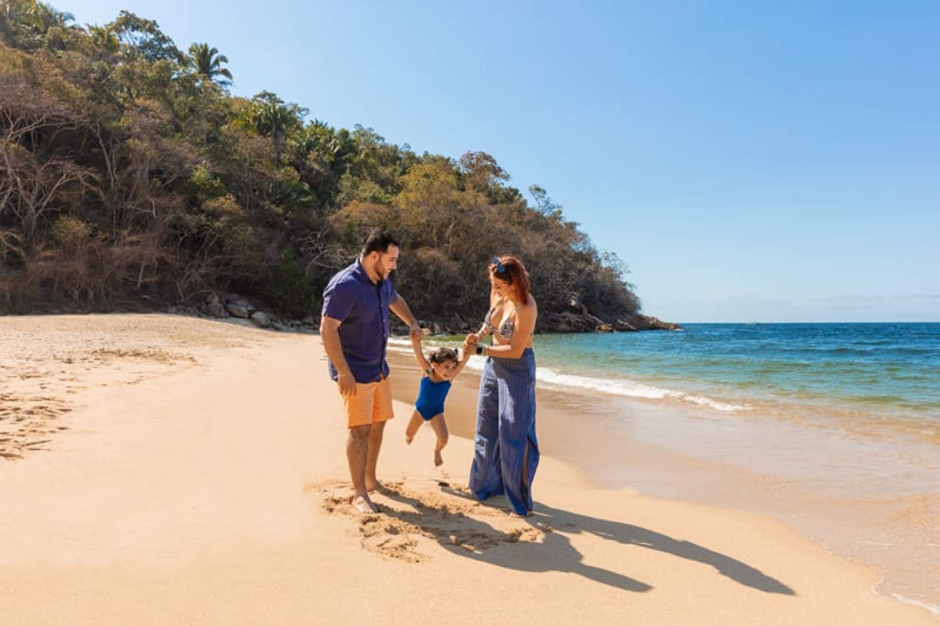 A family enjoys a sunny day on a sandy beach, with parents swinging their child by the hands, blue ocean and green hillside in the background.