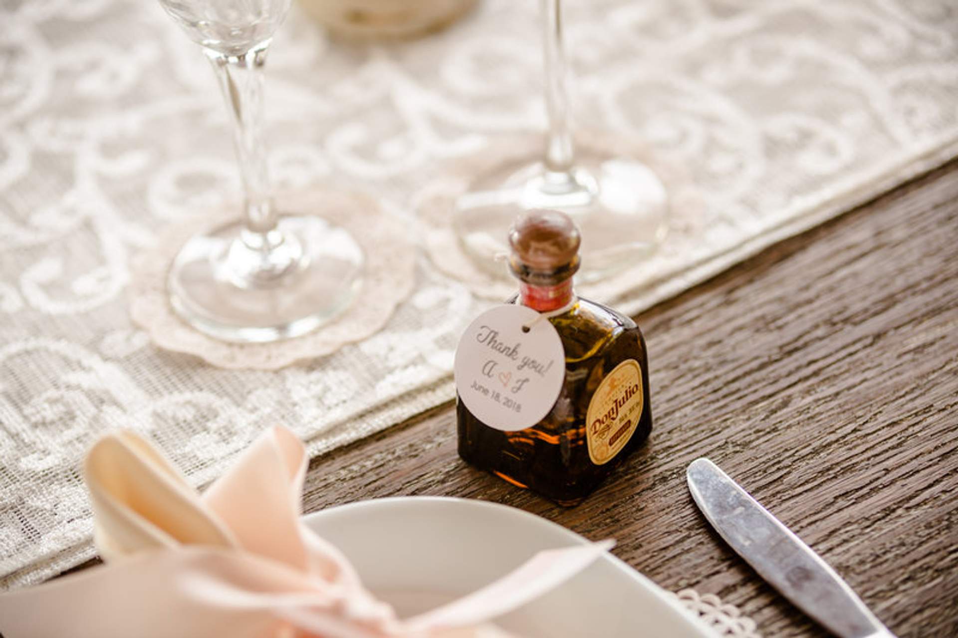 Wedding table setting with a thank-you tag on a mini bottle of tequila, elegant glassware, and a lace table runner.