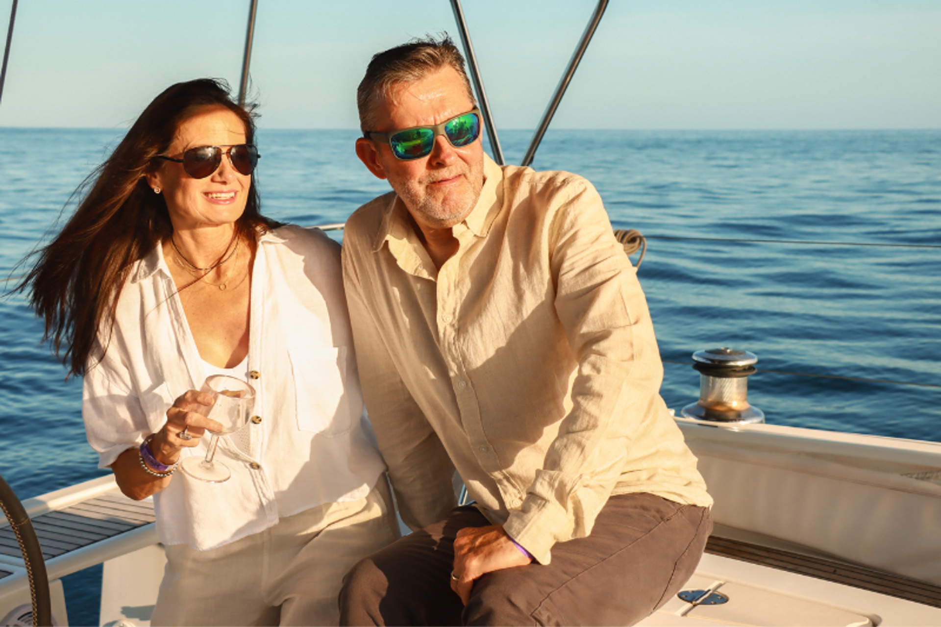 A mature couple enjoying a sailing trip, both wearing sunglasses. The woman is holding a glass of wine and smiling, while the man sits beside her looking out at the ocean.