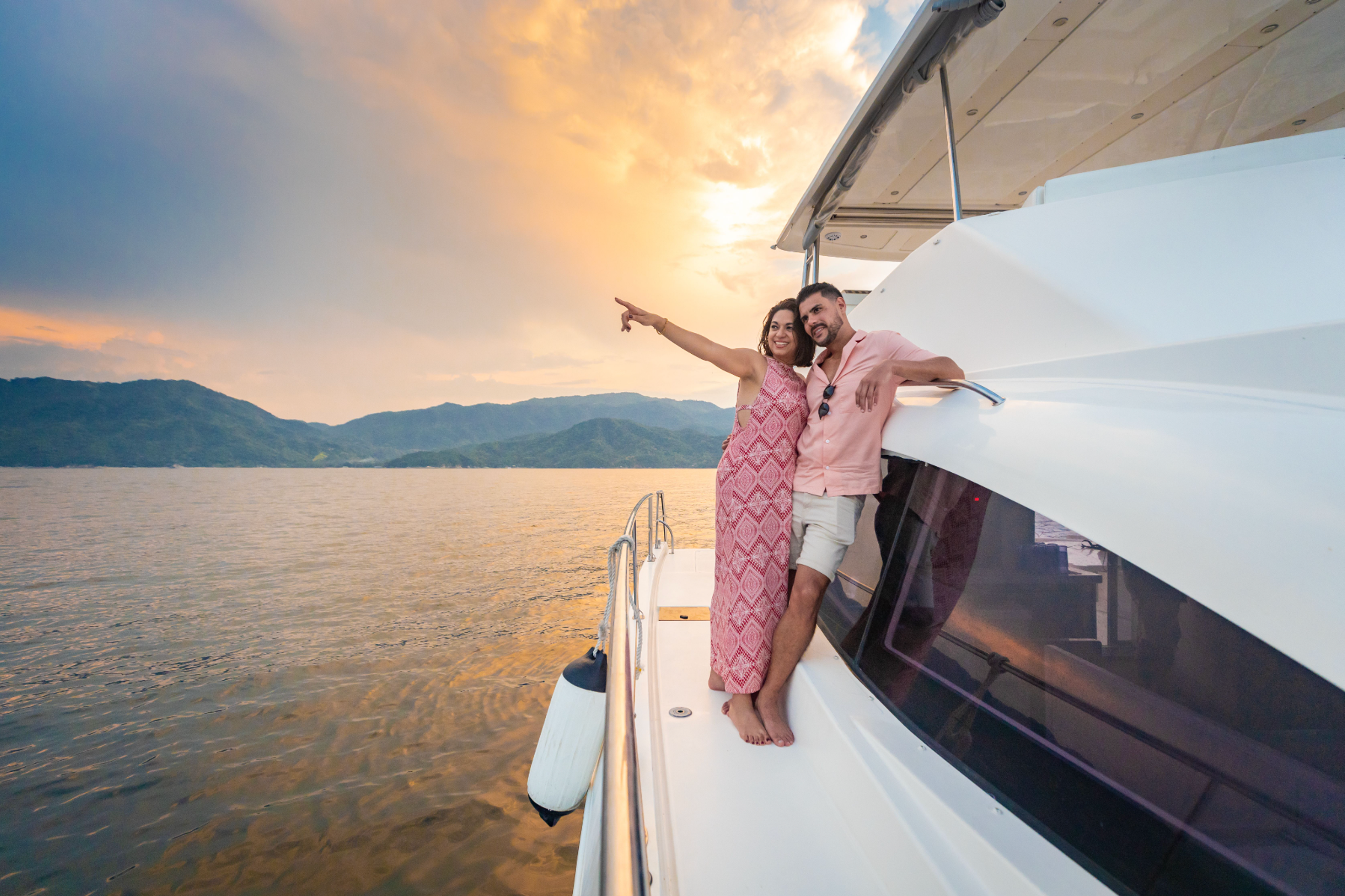A couple enjoying a sunset cruise on a yacht, with the woman pointing towards the scenic mountains in the distance.