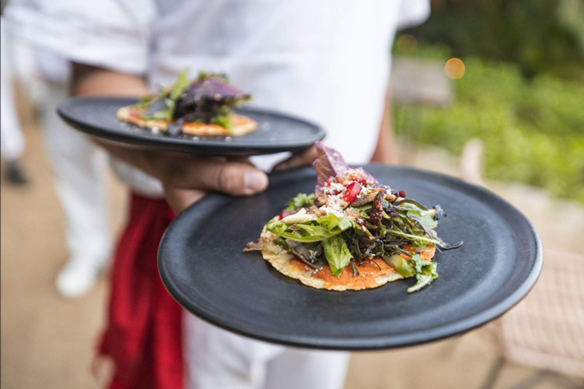 A waiter in white uniform serves two plates of gourmet tacos topped with fresh greens and cheese.