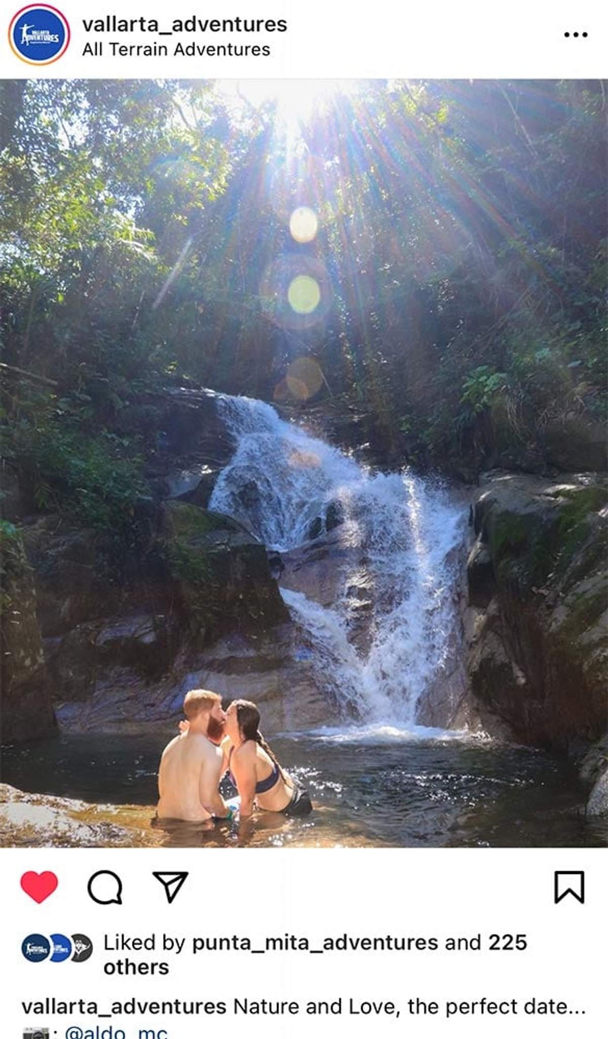 Couple kissing in a natural pool at the base of a waterfall, surrounded by lush greenery and sunlight.
