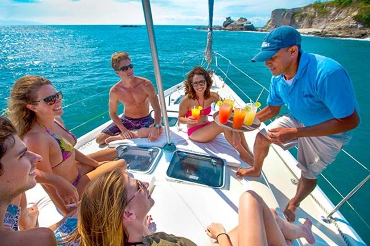 Grupo de amigos en un velero disfrutando de bebidas coloridas servidas por un tripulante con una vista escénica del océano de fondo.