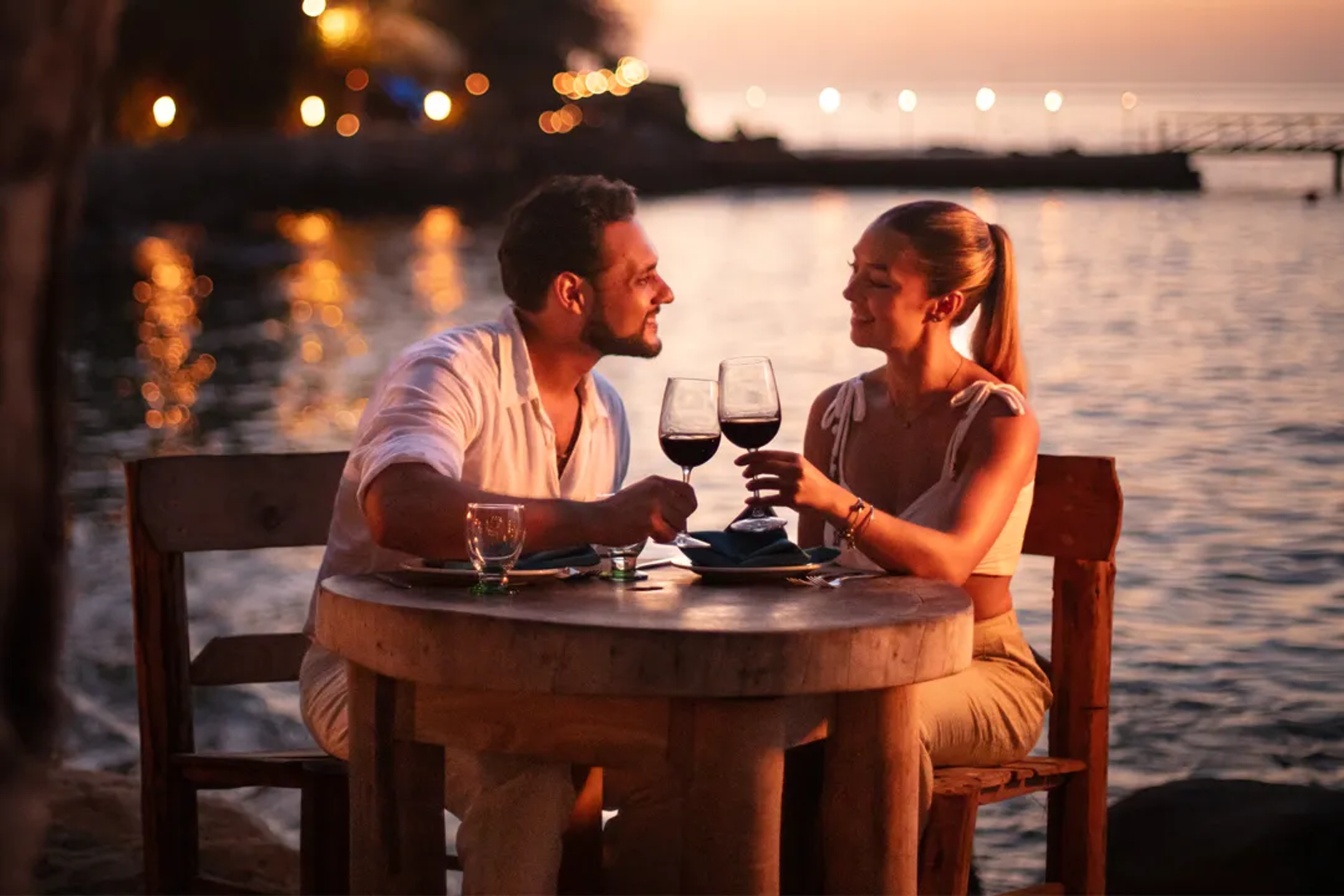 Una pareja brindando con vino durante una cena junto al mar al atardecer.