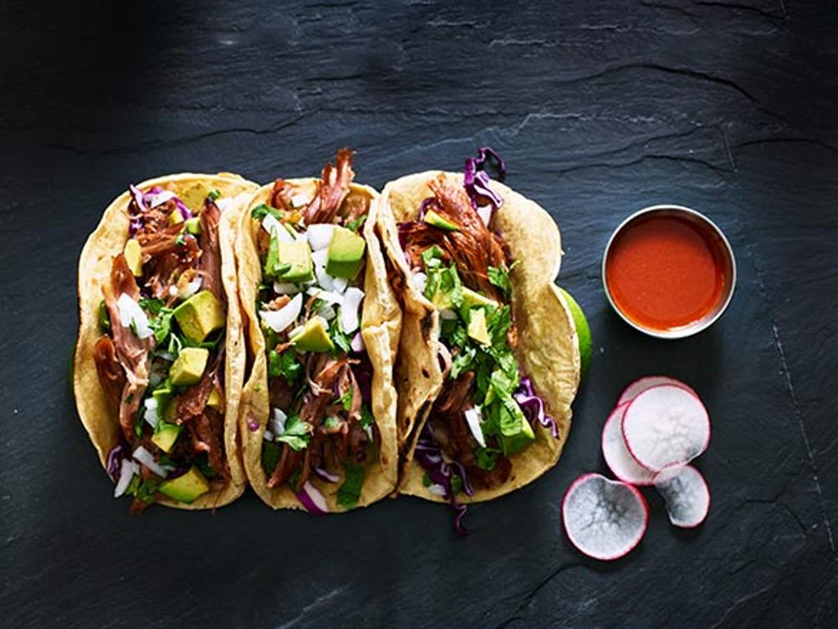 Three delicious tacos filled with meat, avocado, and fresh herbs, served with radish slices and a side of red salsa.