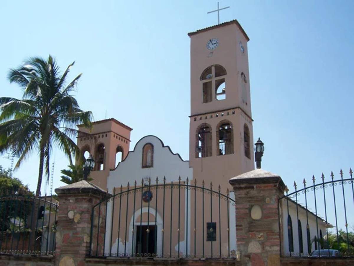A beige church with two bell towers and a cross, surrounded by an iron fence, with a palm tree to the left.