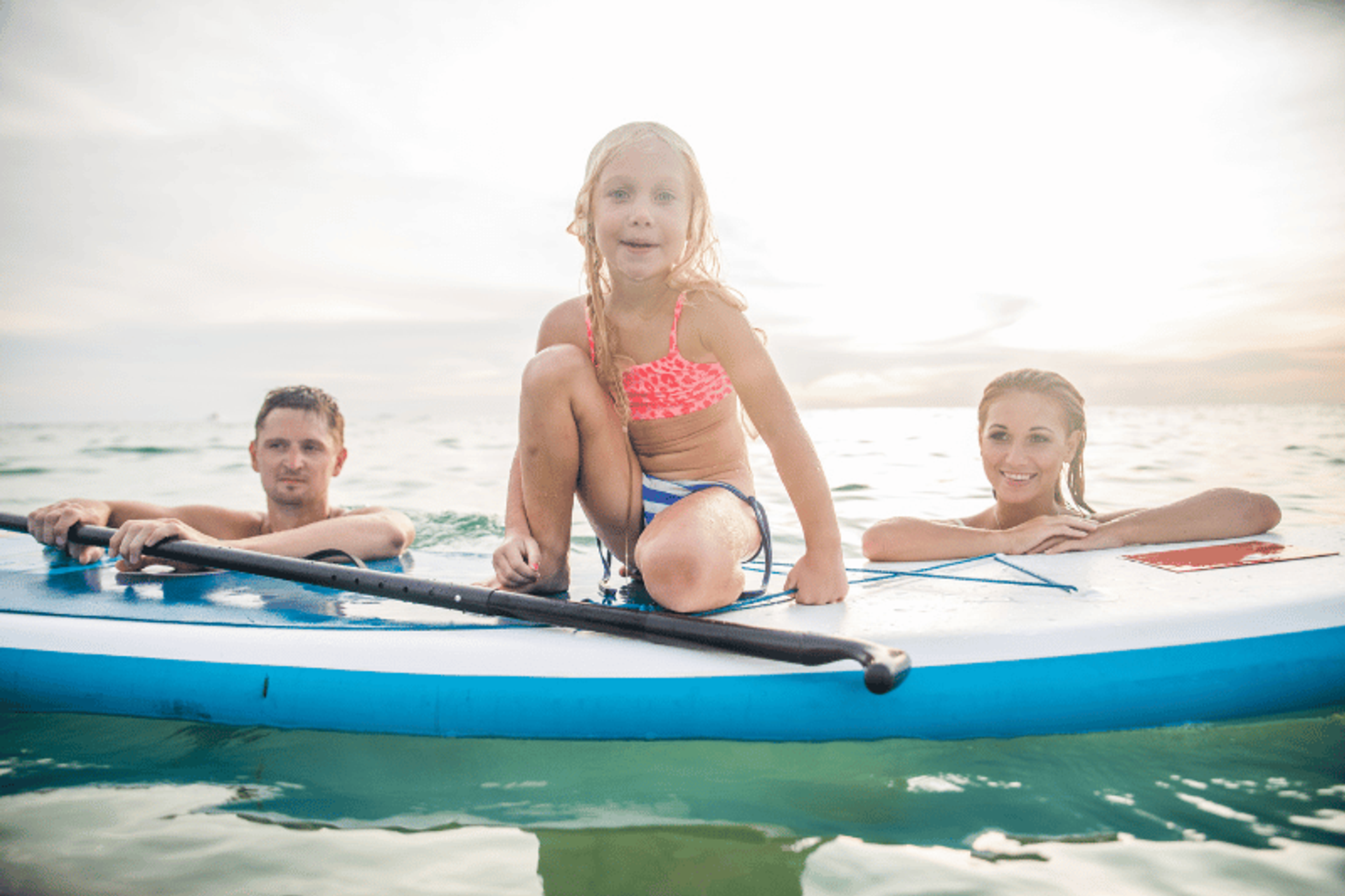 Una familia disfrutando del paddleboarding en el océano. Una niña con un top de bikini rosa se sienta en la tabla de paddle, mientras sus padres nadan a su lado en el agua.