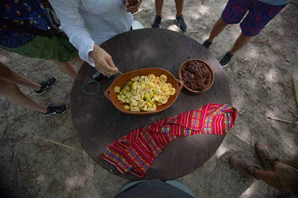 A group of people standing around a round table with traditional dishes, including fried plantains and a colorful woven napkin.