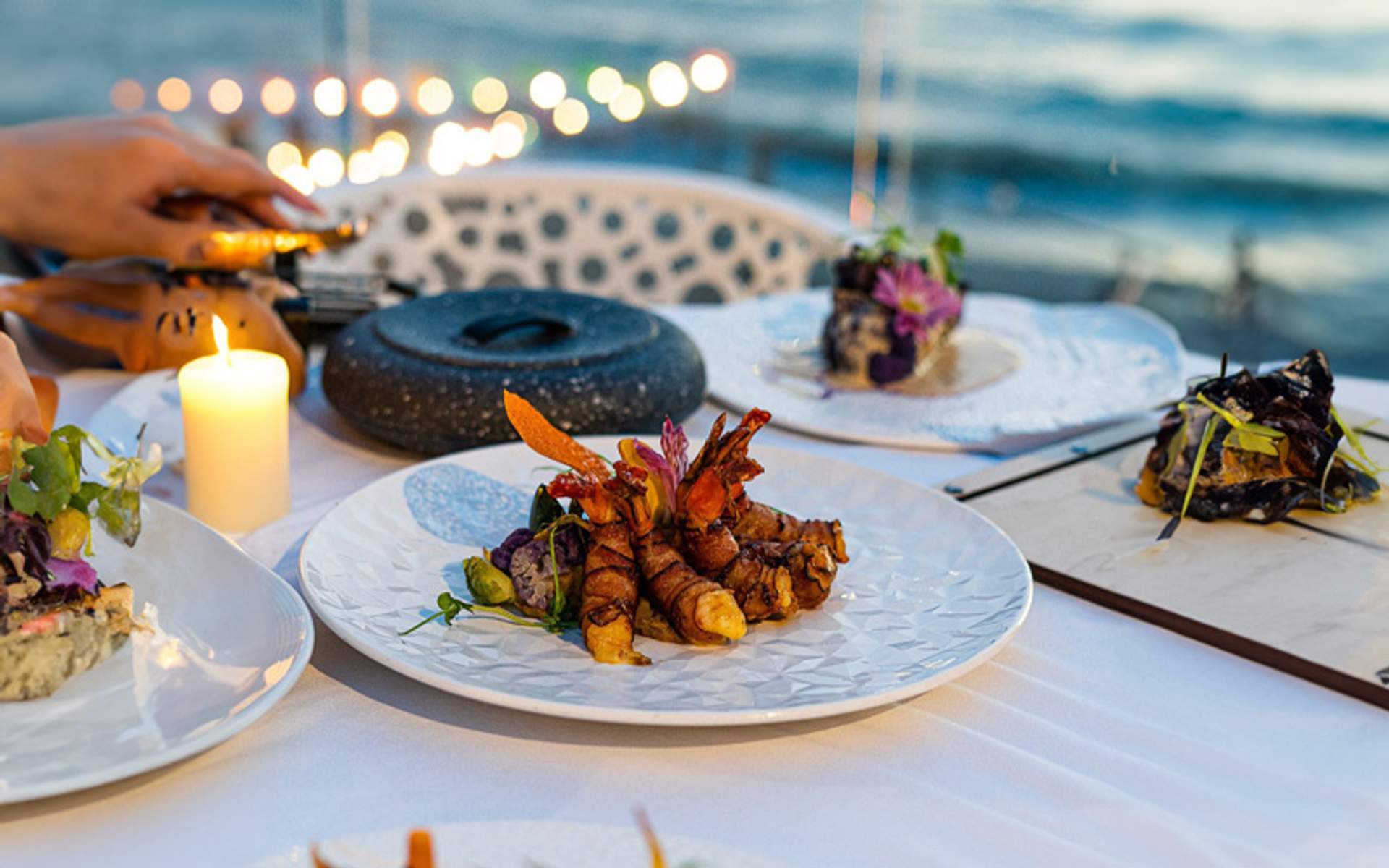 Una cena gourmet al aire libre con platos de mariscos decorados y una vela encendida sobre una mesa blanca, con vista al mar al fondo.