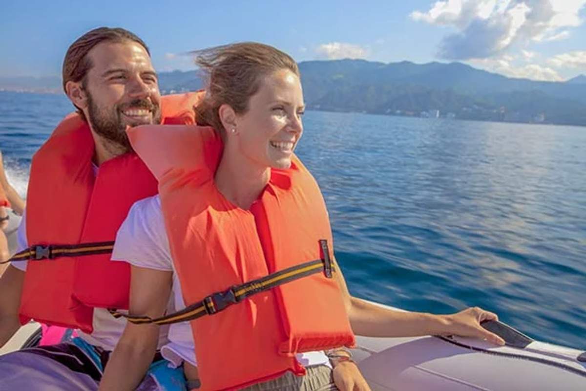 Pareja sonriente con chalecos salvavidas disfrutando de un paseo en bote en aguas azules claras con montañas al fondo.
