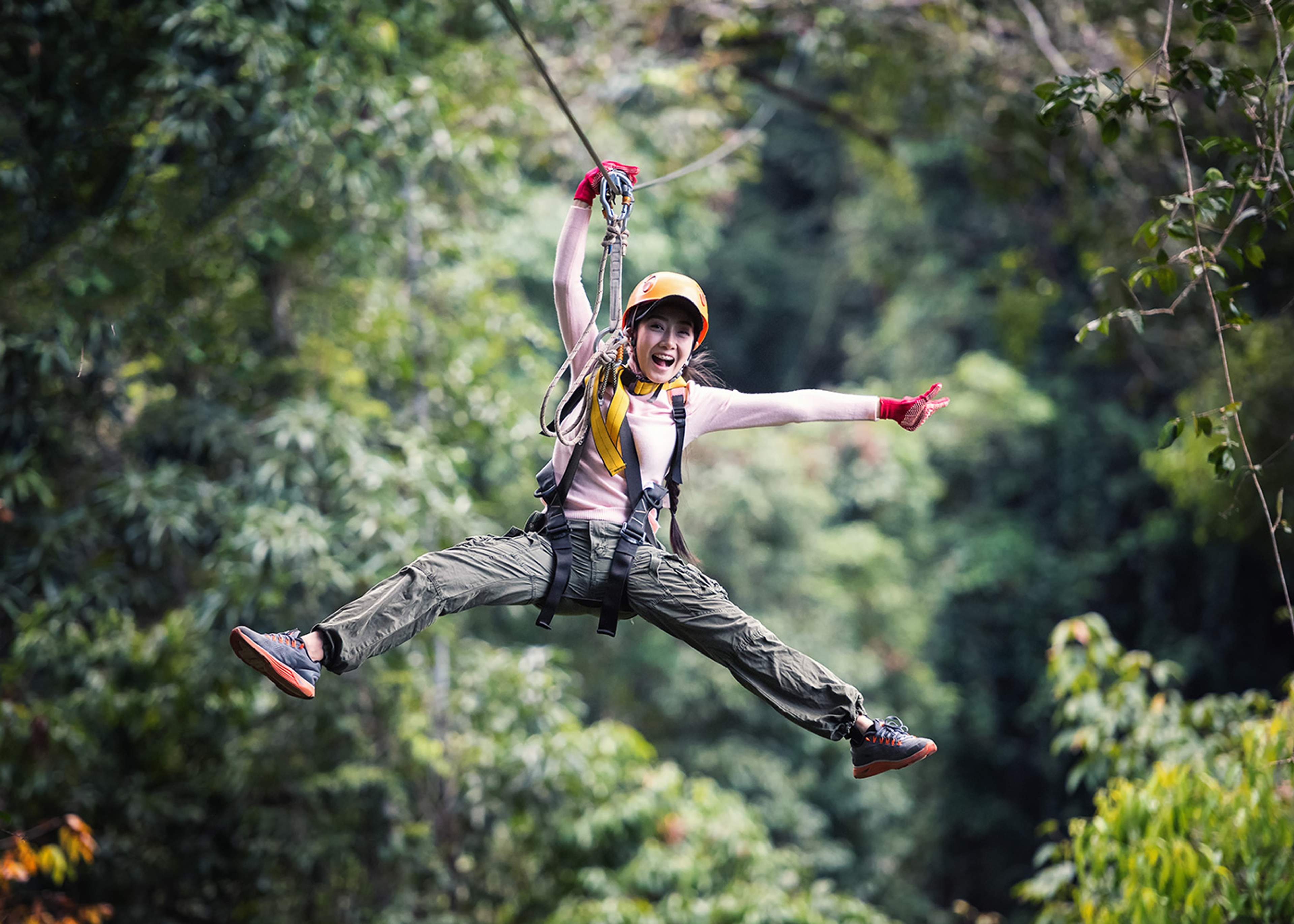 A woman wearing a helmet and harness is ziplining through a forest, with a wide smile and arms outstretched in excitement.
