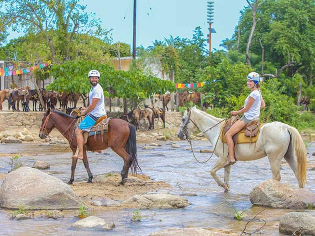 Dos personas disfrutan de un paseo a caballo por un río poco profundo en Quimixto, rodeados de árboles y banderas coloridas.