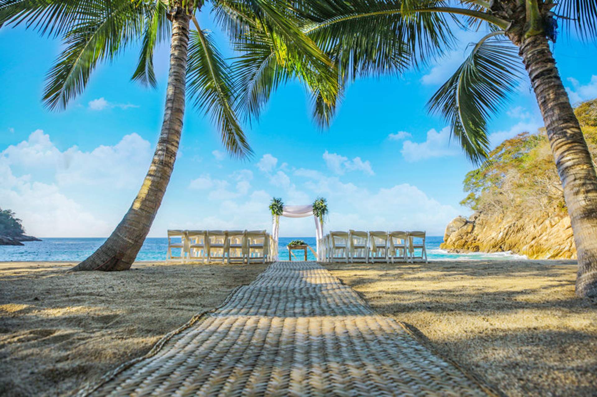 Un hermoso montaje de boda en la playa de Majahuitas, con sillas blancas y un arco floral entre palmeras, frente al océano.
