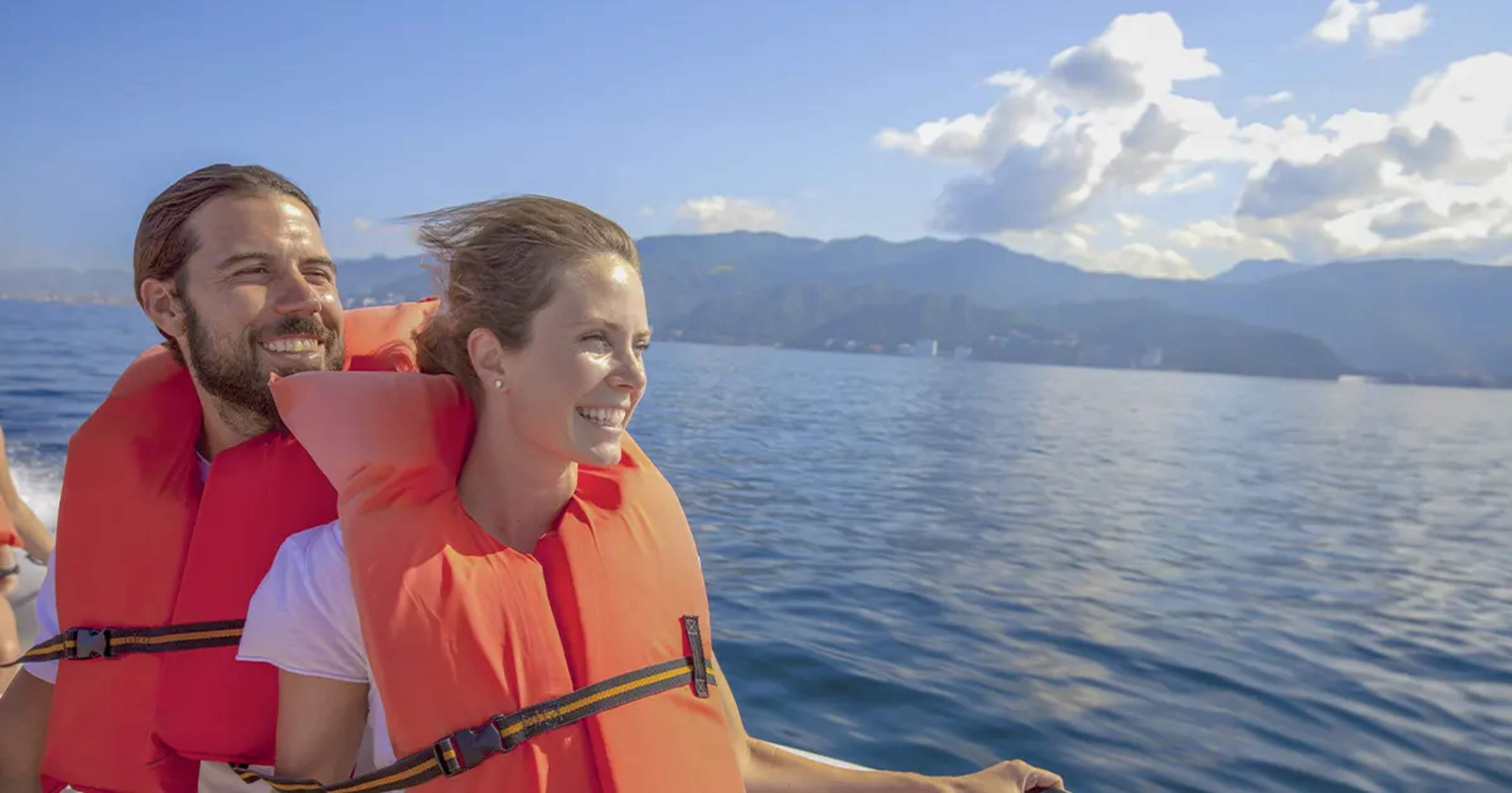A smiling couple wearing life vests enjoying a boat ride on the ocean with mountains in the background under a sunny sky.