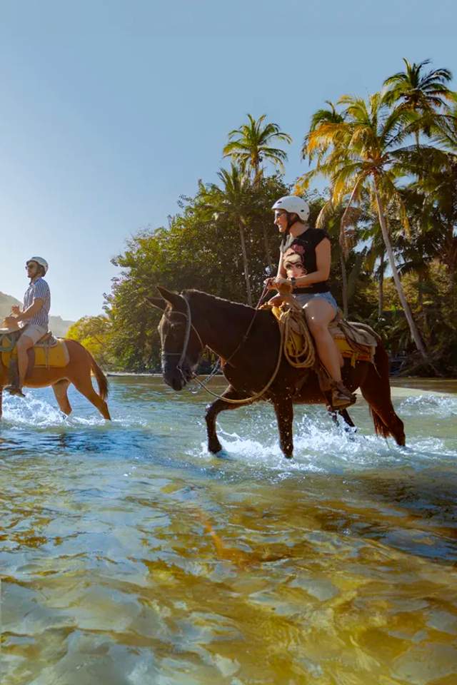 Personas cabalgando a lo largo del río Yelapa con vegetación exuberante y montañas al fondo.