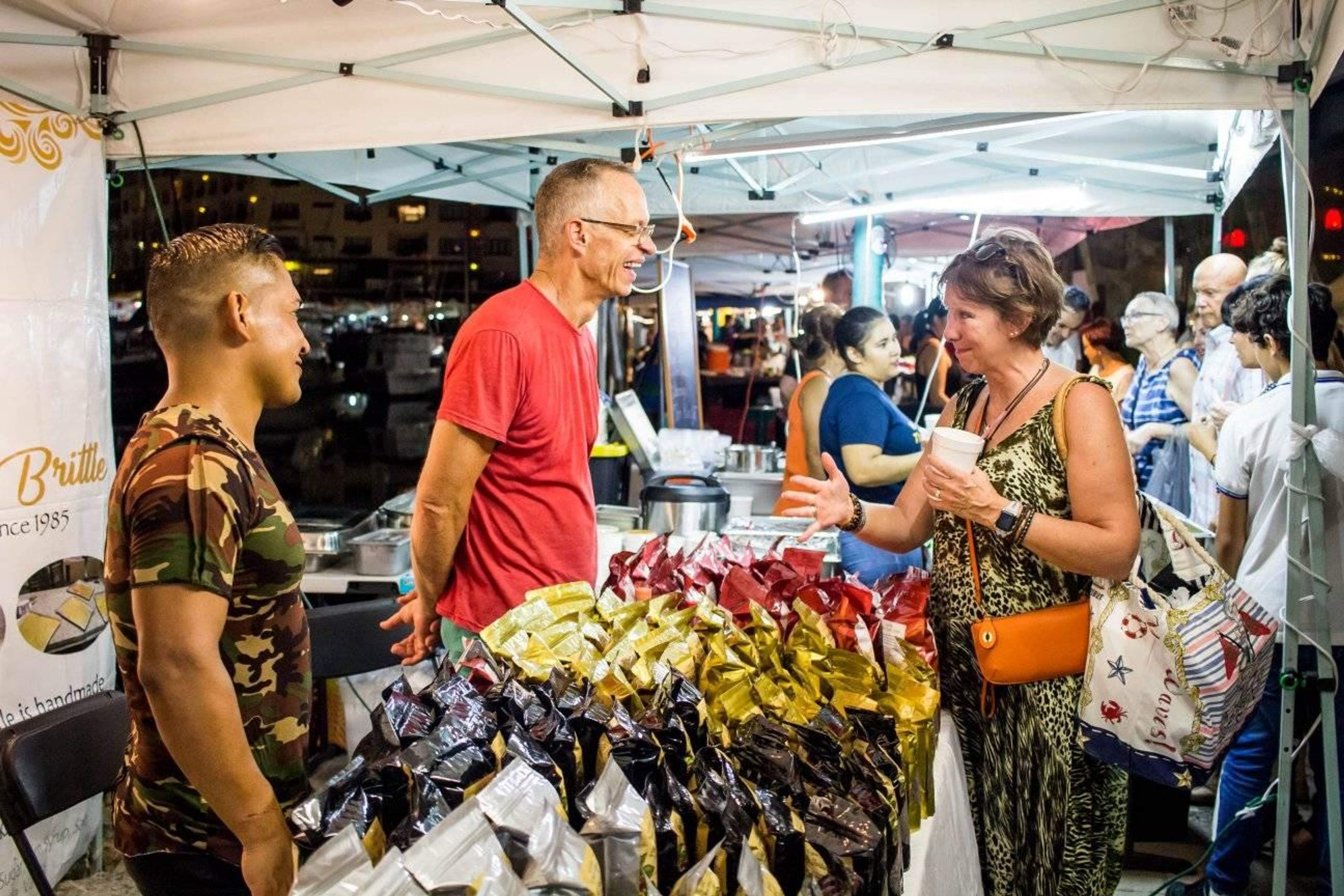 Personas interactuando en un puesto de mercado bajo una carpa por la noche. Un vendedor con una camiseta roja y otro con una camiseta de camuflaje están hablando y sonriendo con una mujer que sostiene una bebida. El puesto está lleno de bolsas de varios productos.