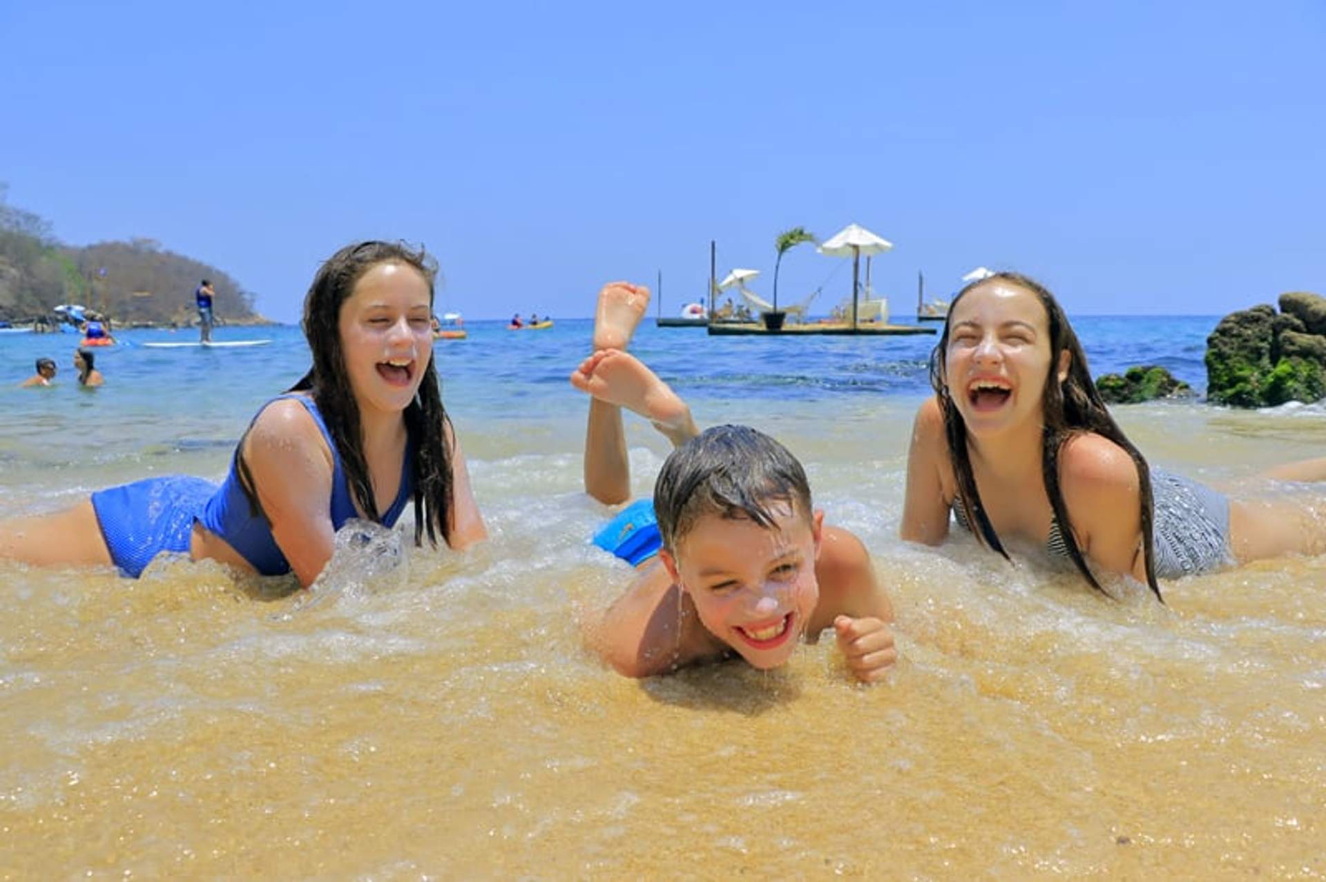 Tres niños se divierten en la orilla de la playa, riendo mientras juegan en el agua con un muelle y sombrillas al fondo.