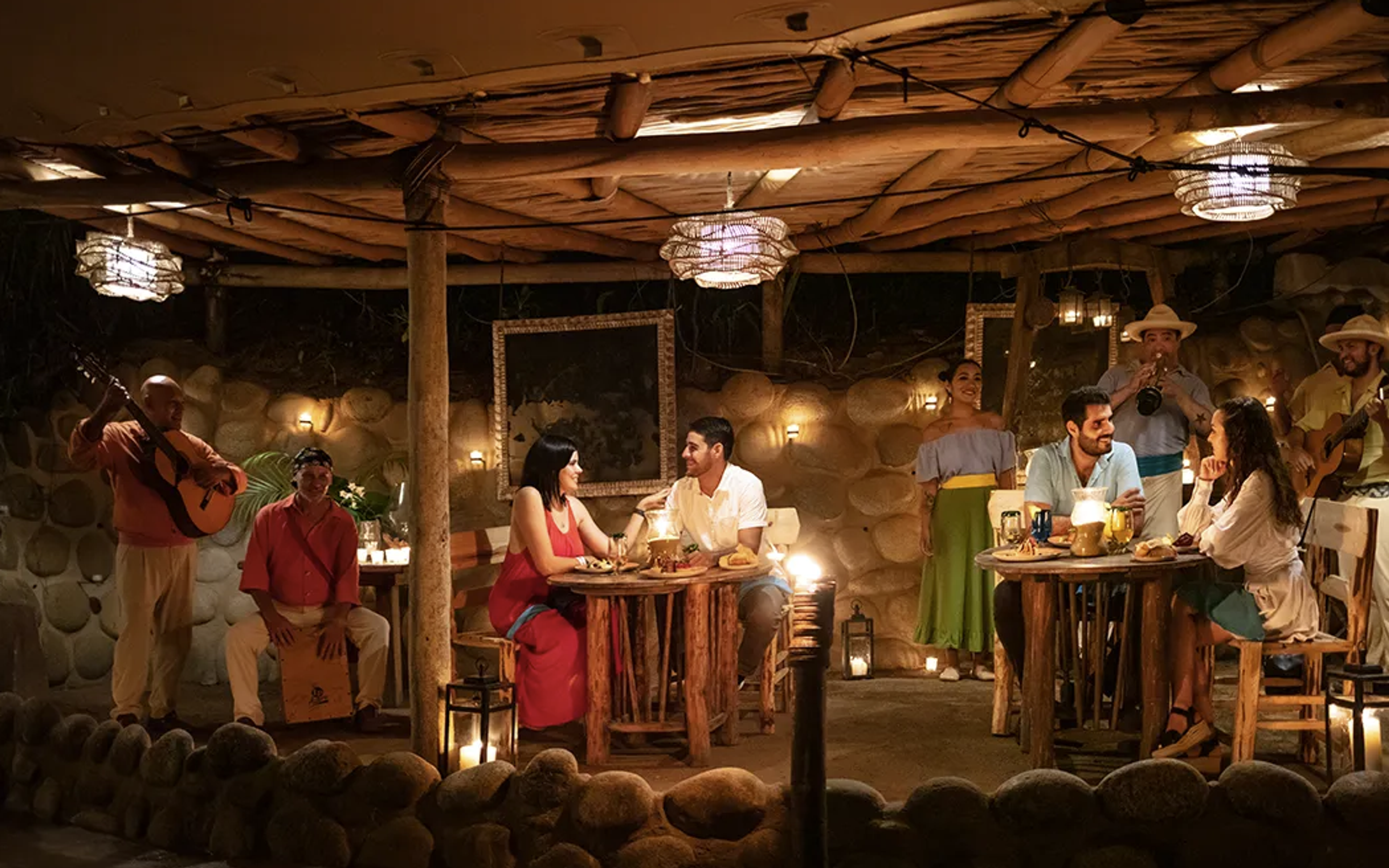 Una cena al aire libre con dos parejas disfrutando de comida y música en vivo, bajo una iluminación cálida y acogedora.